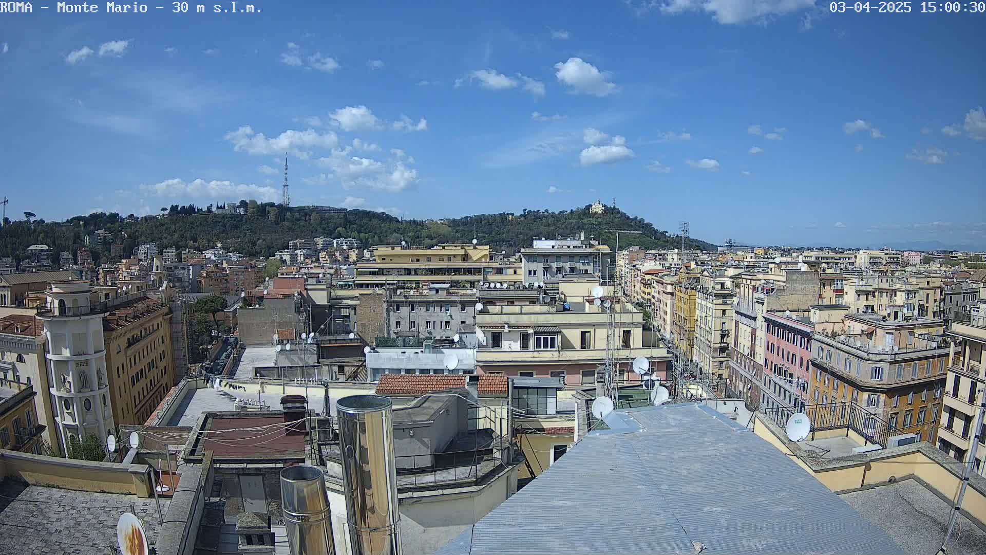 A mostly sunny day reveals a cityscape of densely packed buildings, with a hill covered in green vegetation in the background.