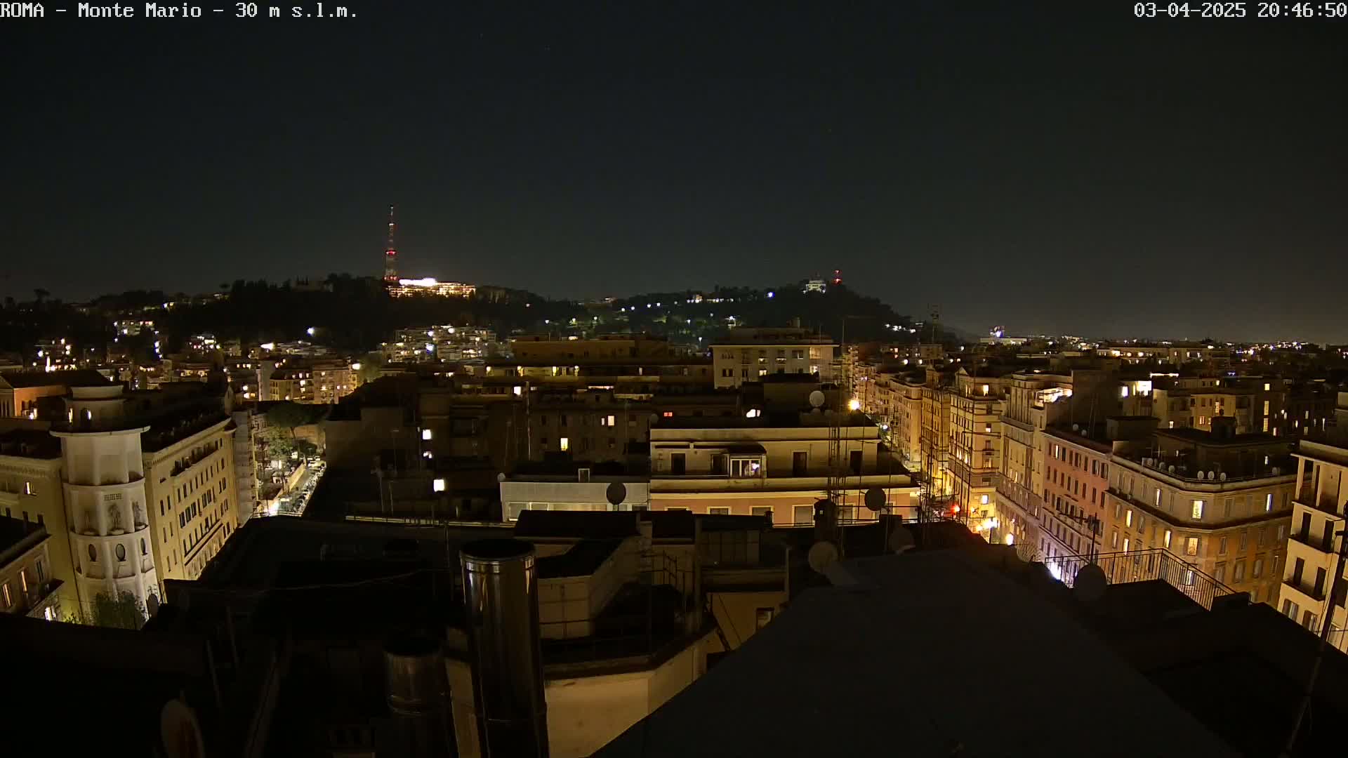 A nighttime aerial view of a city with illuminated buildings extending to a hilltop in the distance under a clear, dark sky.
