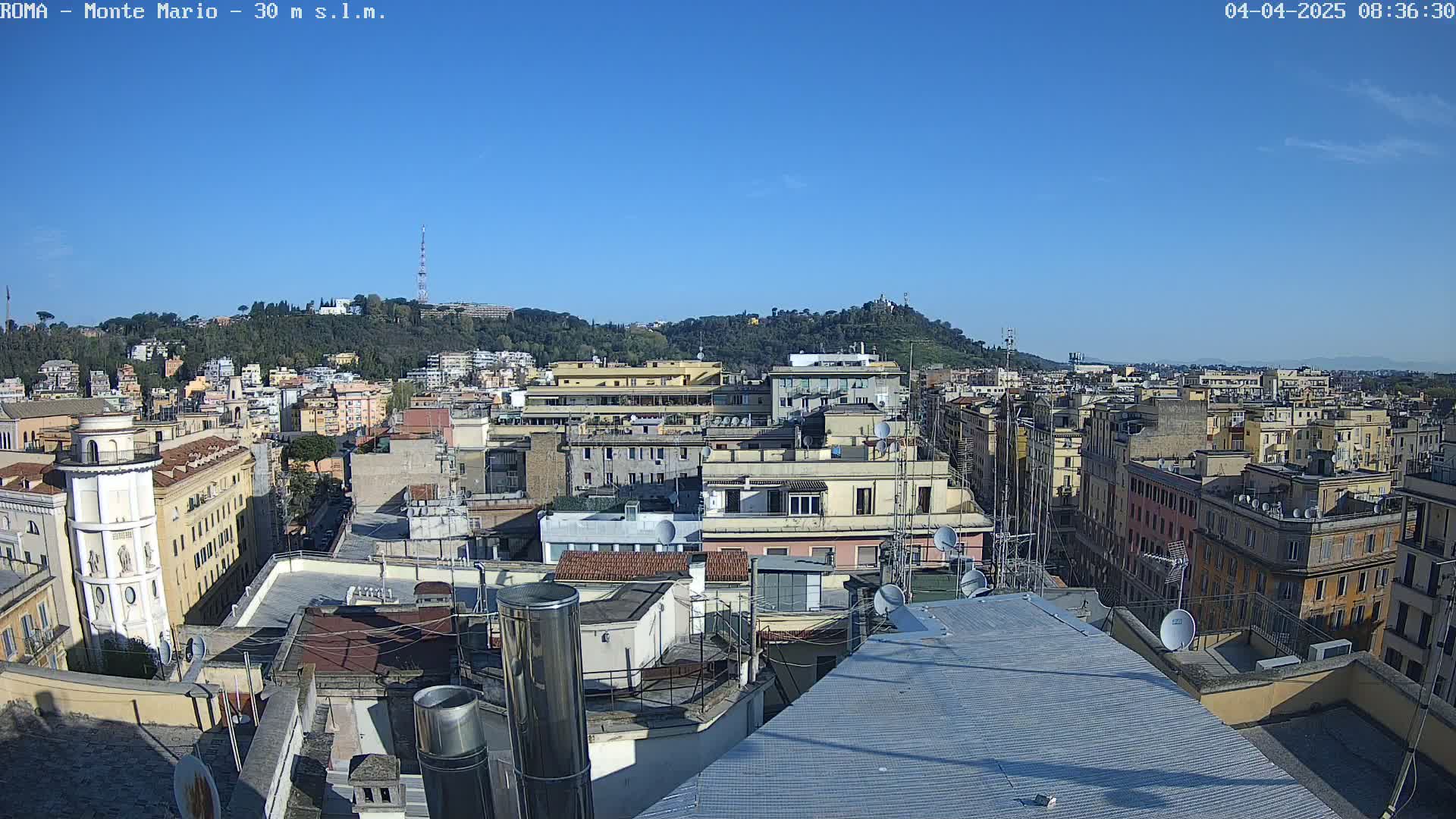A panoramic view of a city on a clear, sunny day shows many rooftops, buildings, and a hill in the distance.