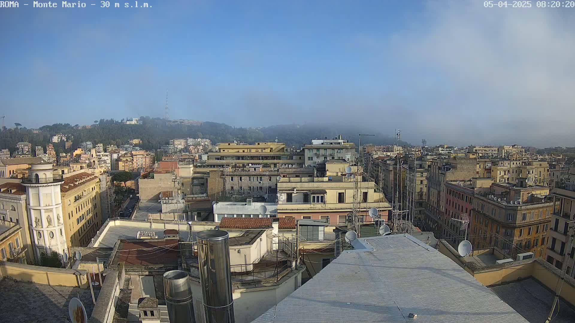 A hazy, clear-sky view from a rooftop overlooking a dense cityscape of multi-story buildings, extending to a distant hill in the background.