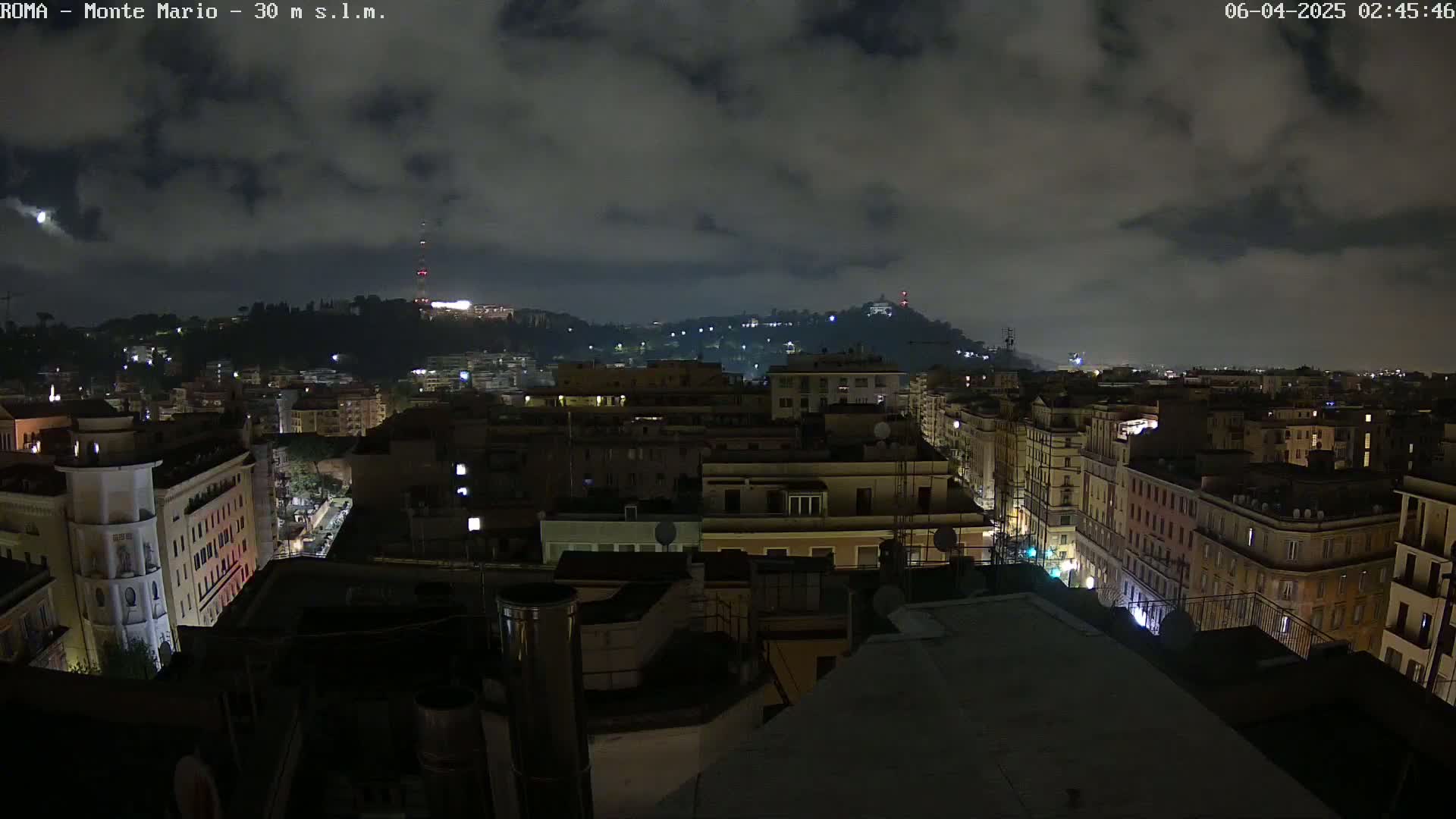 A nighttime cityscape with a cloudy sky shows numerous buildings illuminated with some lights on a street below, extending into a hill in the distance.