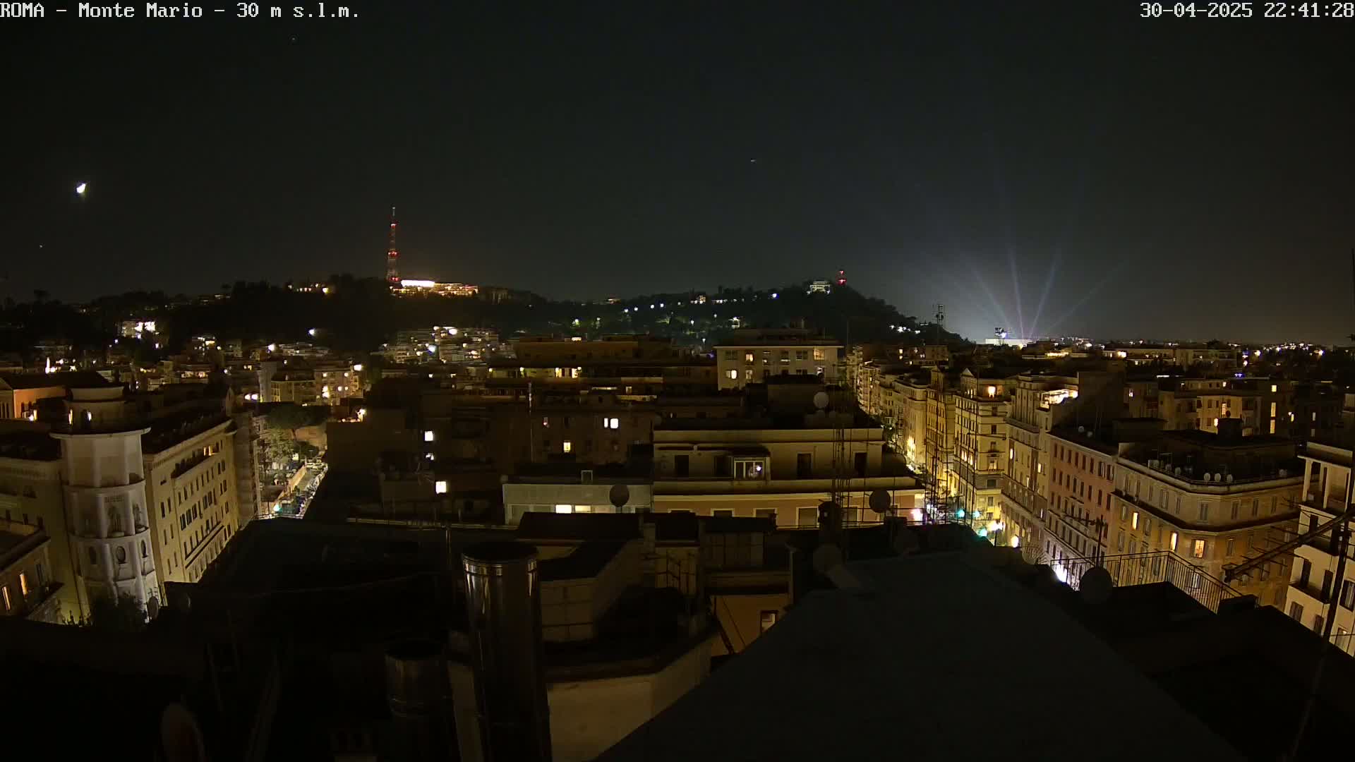 A nighttime view of a city illuminated by lights, with a hill in the background under a clear, dark sky.