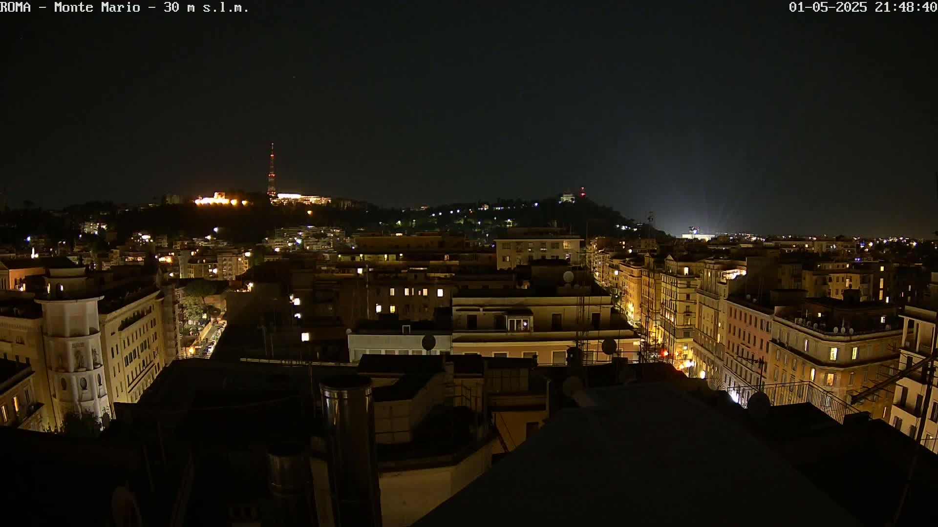 A nighttime view of a city, showing illuminated buildings and streets extending to a hilltop in the distance under a clear, dark sky.
