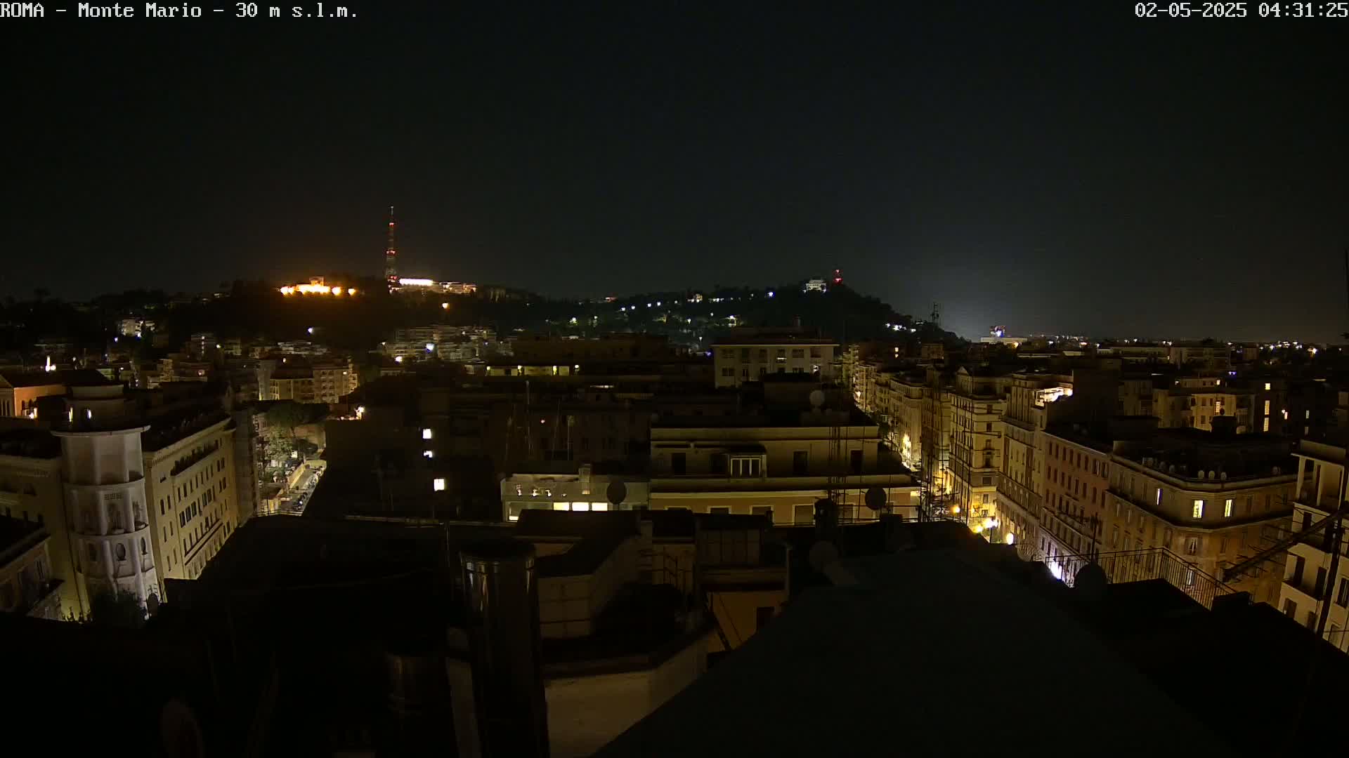 A nighttime, high-angle view of a city's cityscape, showing numerous buildings with illuminated windows, and hills in the distance under a dark, clear sky.