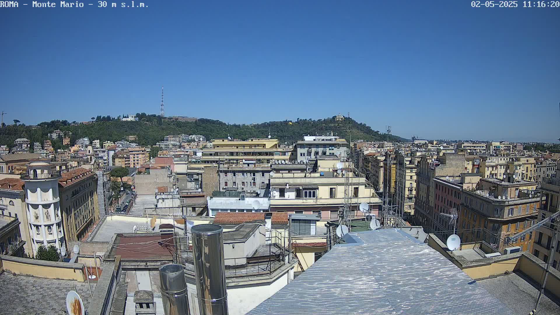 A rooftop view of a dense city on a clear, sunny day, showing numerous buildings extending to a hill in the distance.