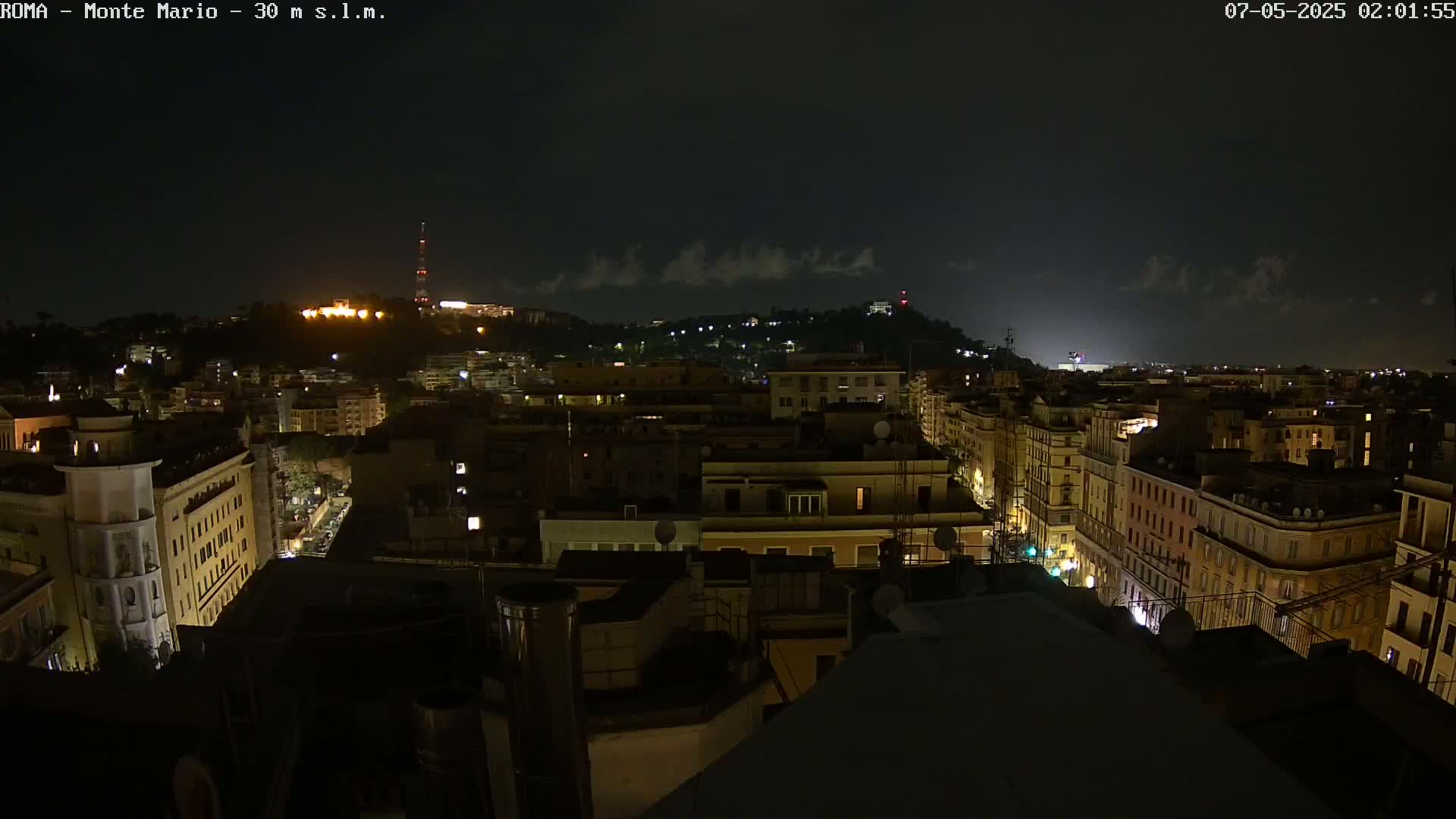 A nighttime, high-angle view of a city with dimly lit buildings under a mostly cloudy sky.