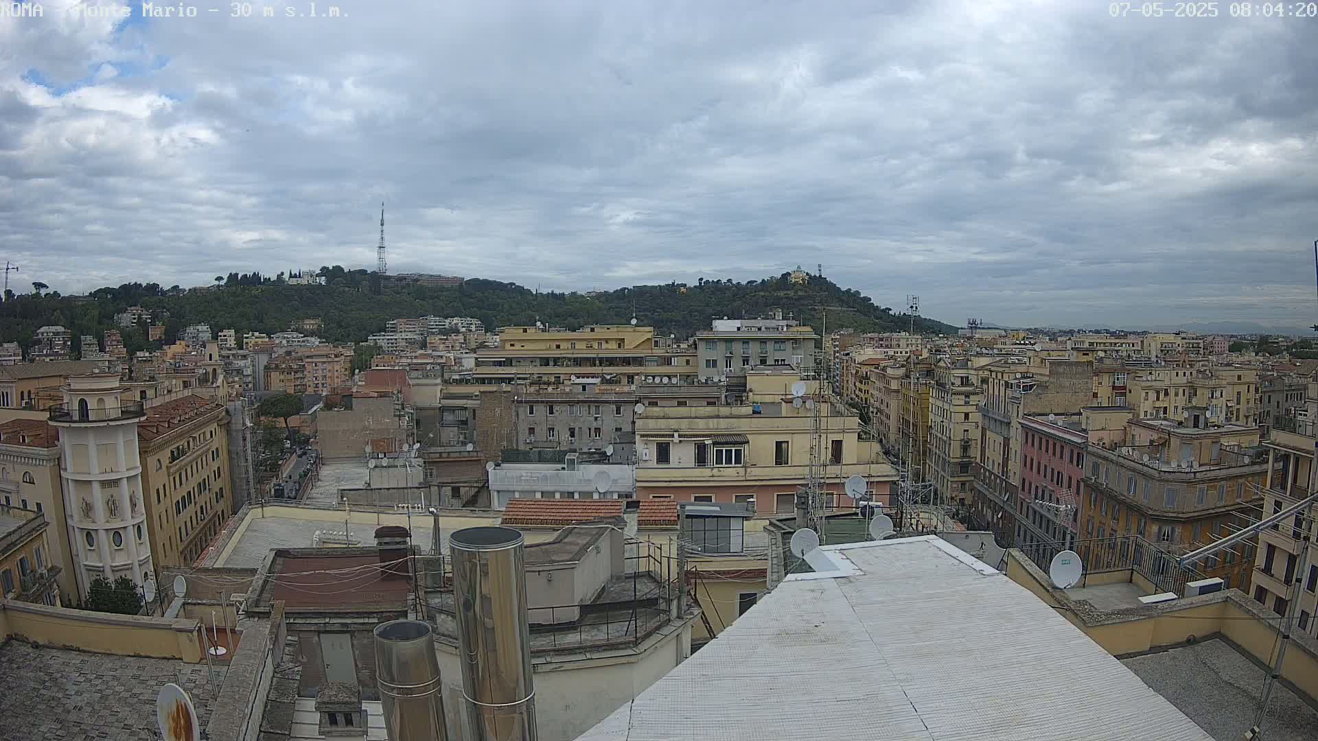 A view from a rooftop shows a cityscape of closely packed buildings under a cloudy sky, with a hill in the background.