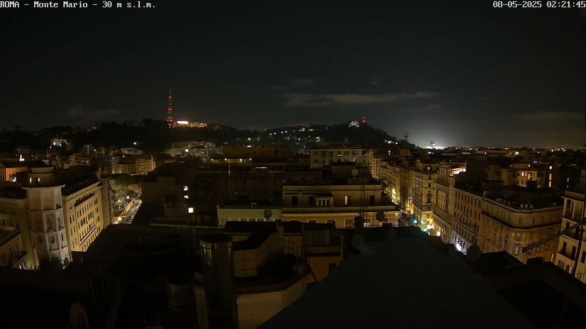 A nighttime cityscape shows a hilly area with buildings lit up under a mostly clear, dark sky.