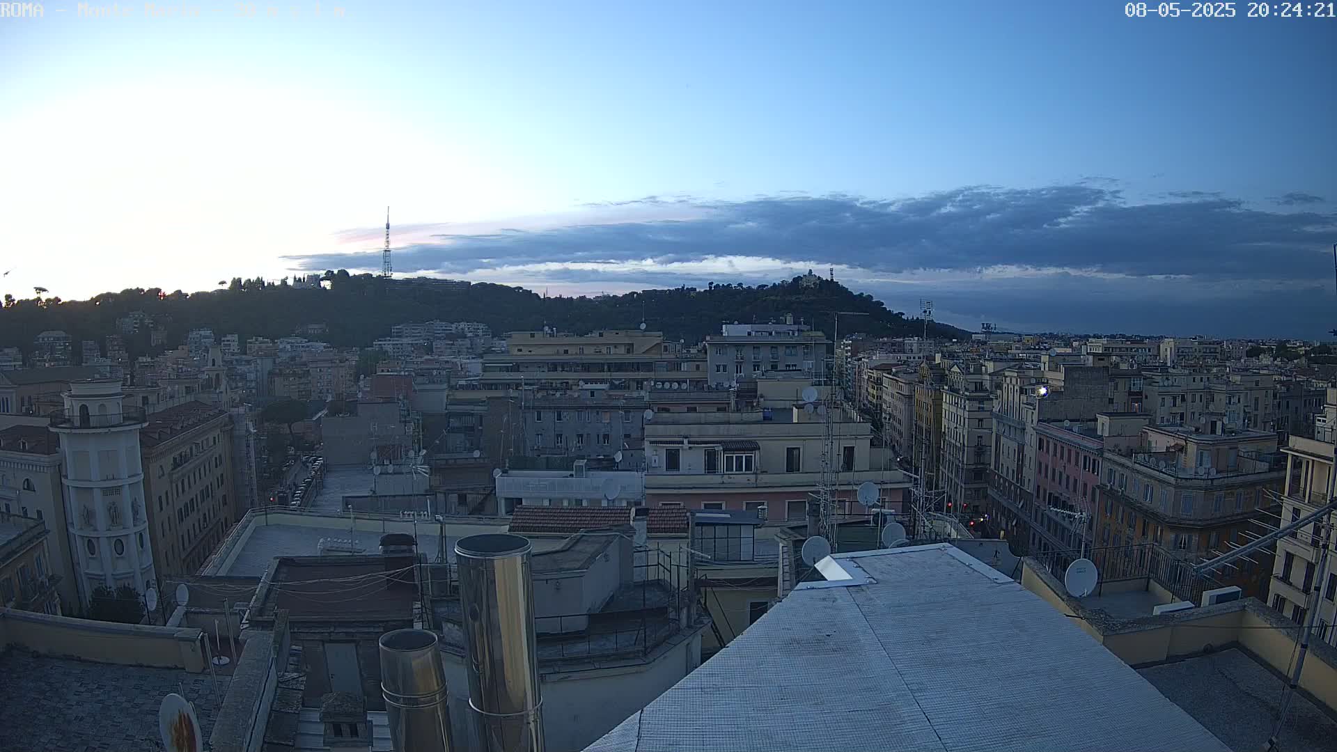 A cityscape at dusk under a partly cloudy sky, viewed from a rooftop, shows many buildings stretching to a hill in the distance.