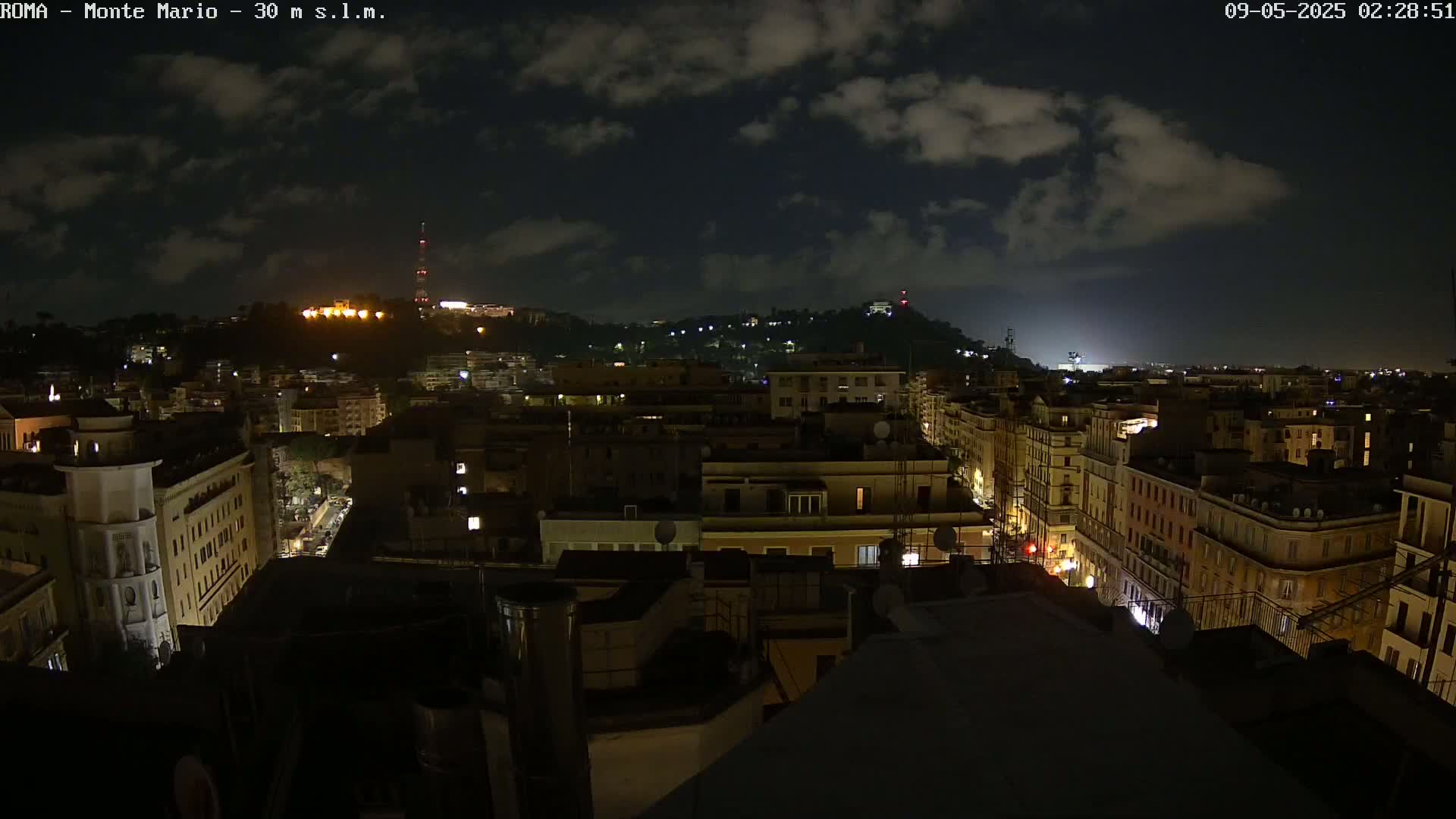 A nighttime view of a city, under a partly cloudy sky, shows numerous buildings illuminated with lights against a dark background.