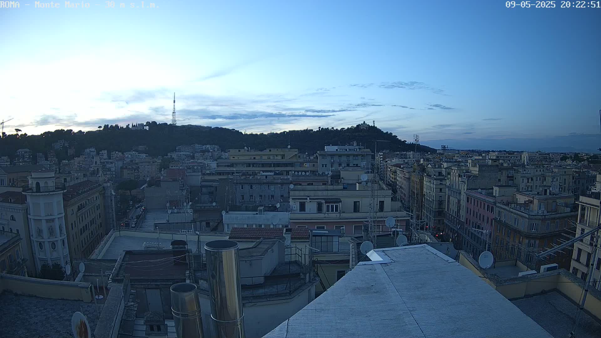 A city skyline at dusk, under a mostly clear sky, shows many buildings, rooftops, and a hill in the distance.