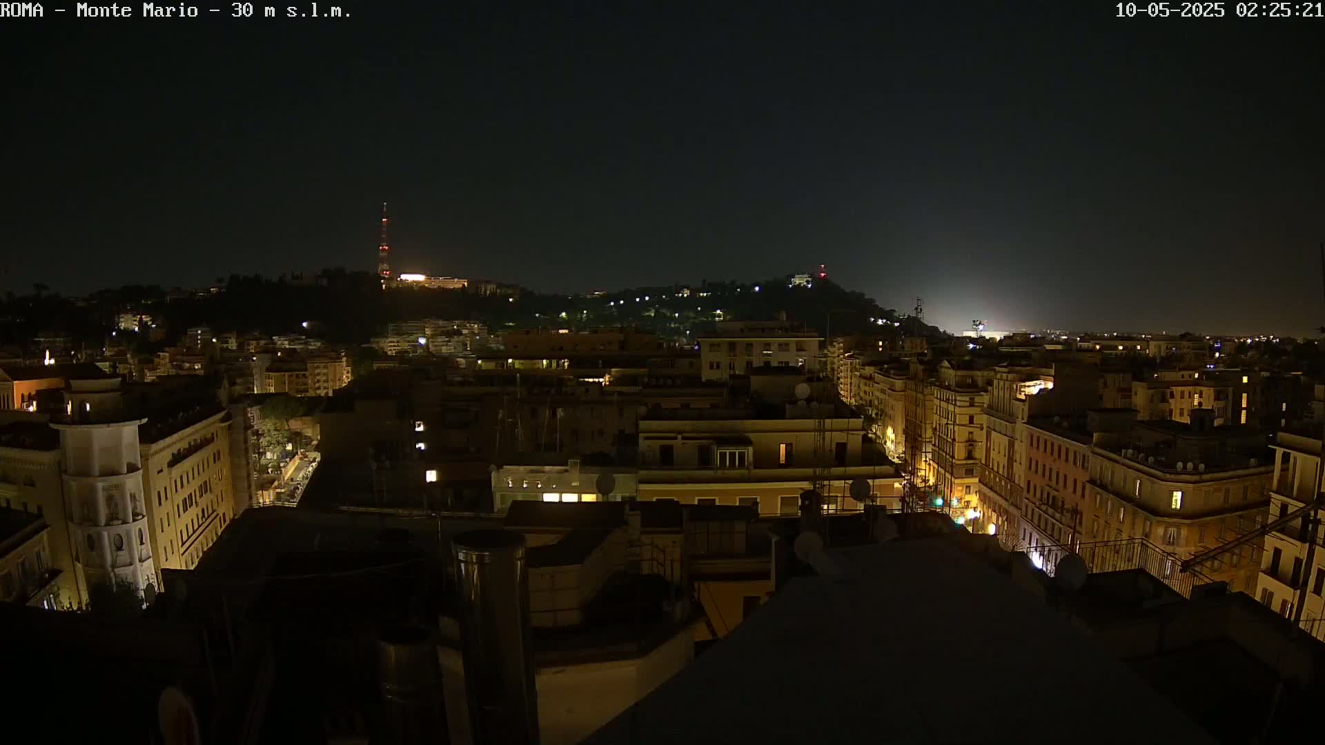 A nighttime view of a city with illuminated buildings and streets, overlooking a hill in the distance under a dark, clear sky.