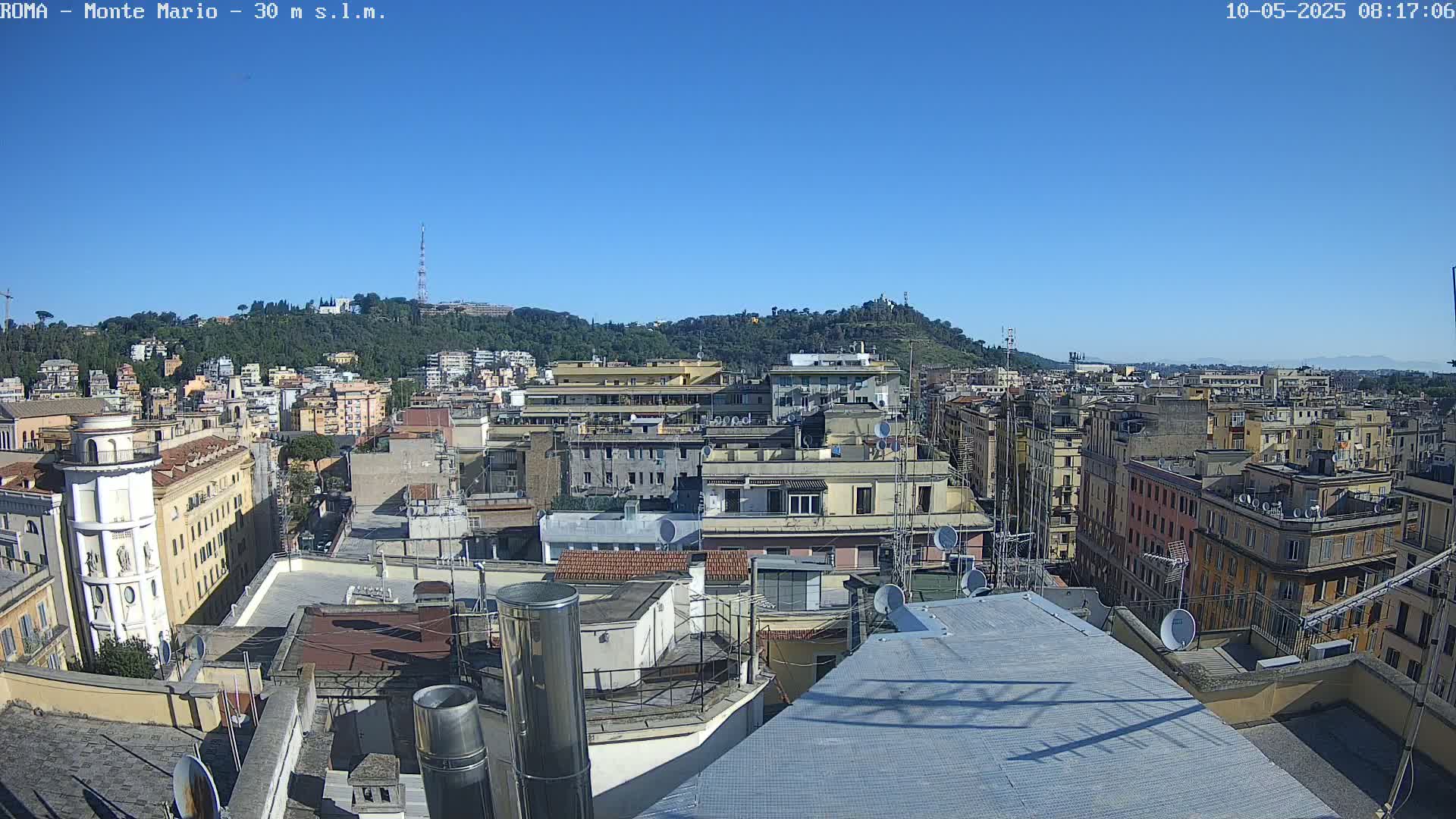 A clear, sunny day reveals a sprawling cityscape of numerous buildings, rooftops, and satellite dishes, with a hill covered in greenery in the background.