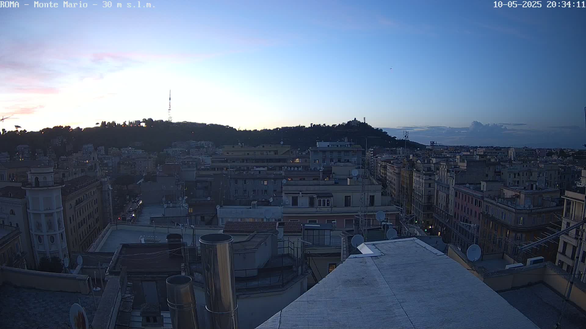 A twilight view of a city skyline, showing many buildings with rooftops and a hill in the background under a mostly clear sky.
