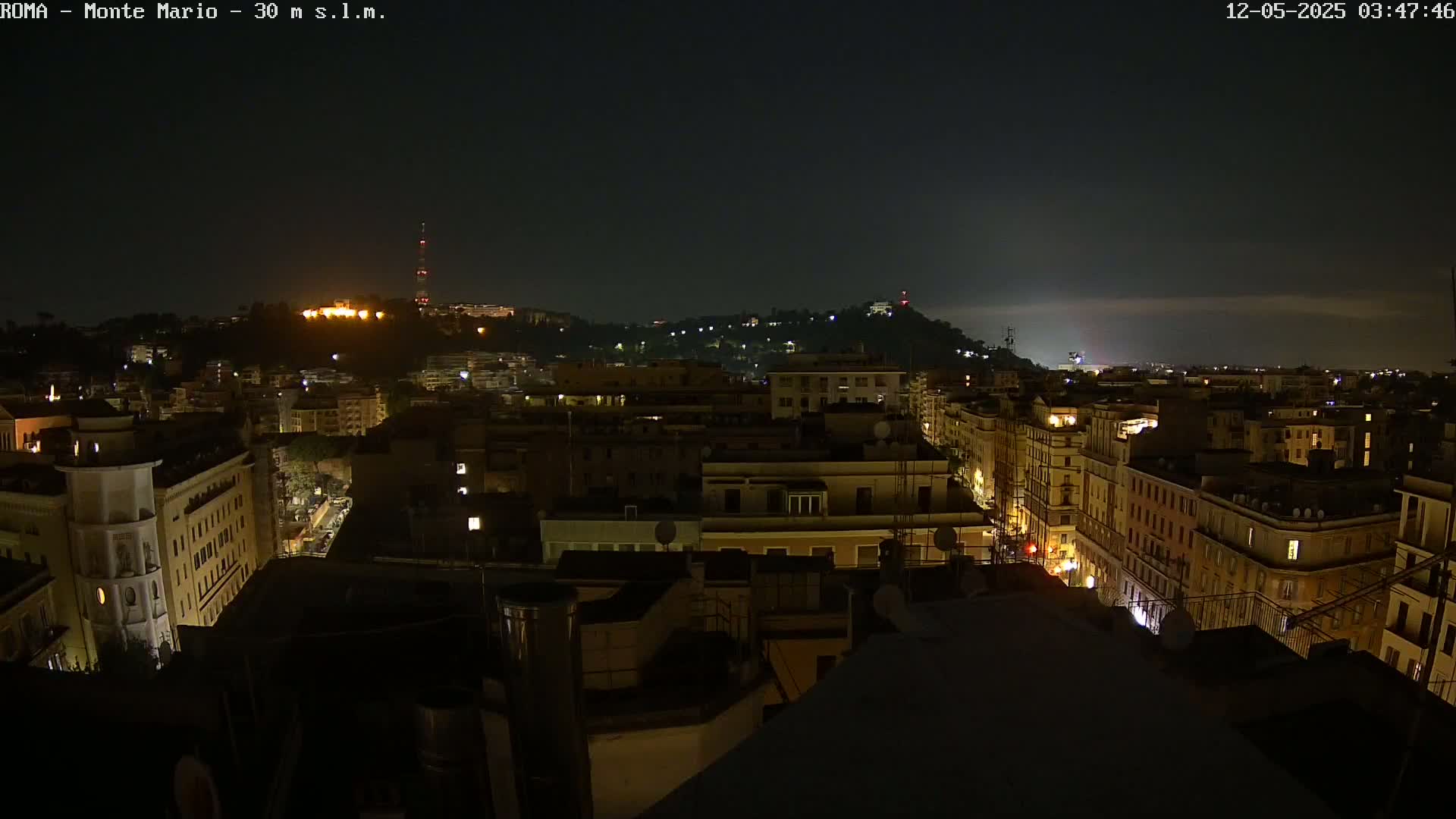 A nighttime view of a city, showing a hilly cityscape with illuminated buildings and streets under a dark, mostly clear sky.