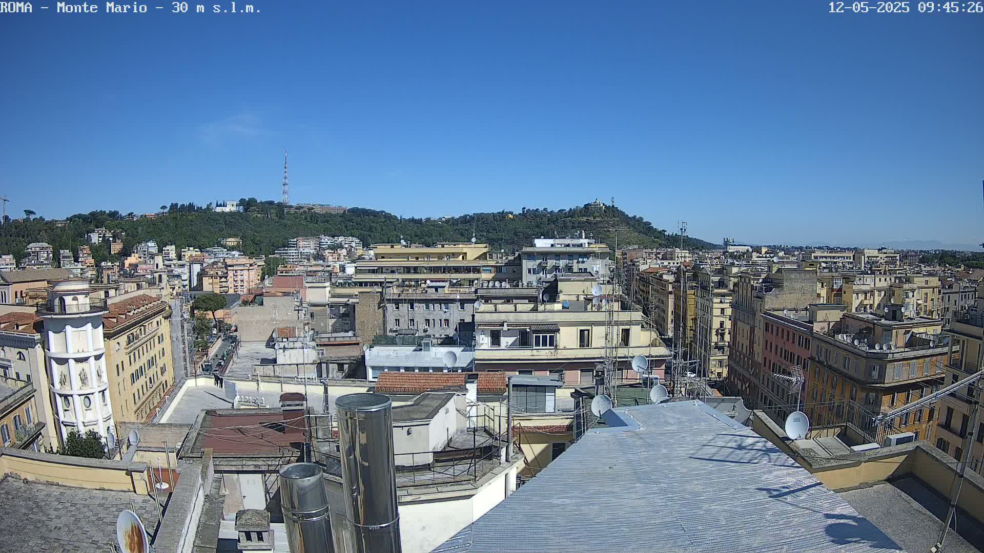 A high-angle view on a clear, sunny day shows numerous rooftops and buildings densely packed together, extending to a hill in the distance.
