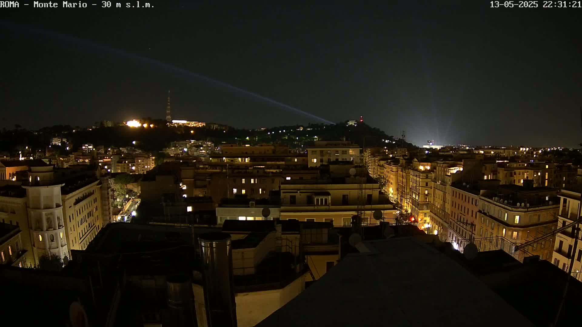 A nighttime view of a city, illuminated by streetlights and building lights, with a hill in the background faintly lit and beams of light projecting into the sky.