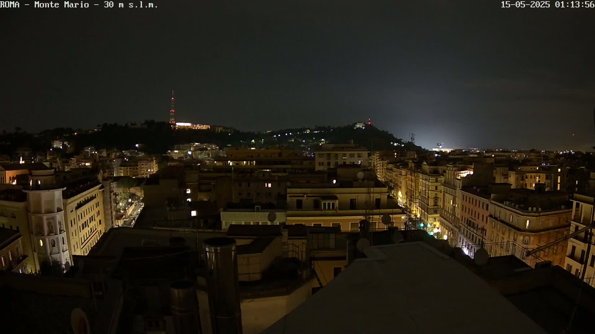 A nighttime aerial view of a city with dimly lit buildings and a hill in the background under a dark sky.