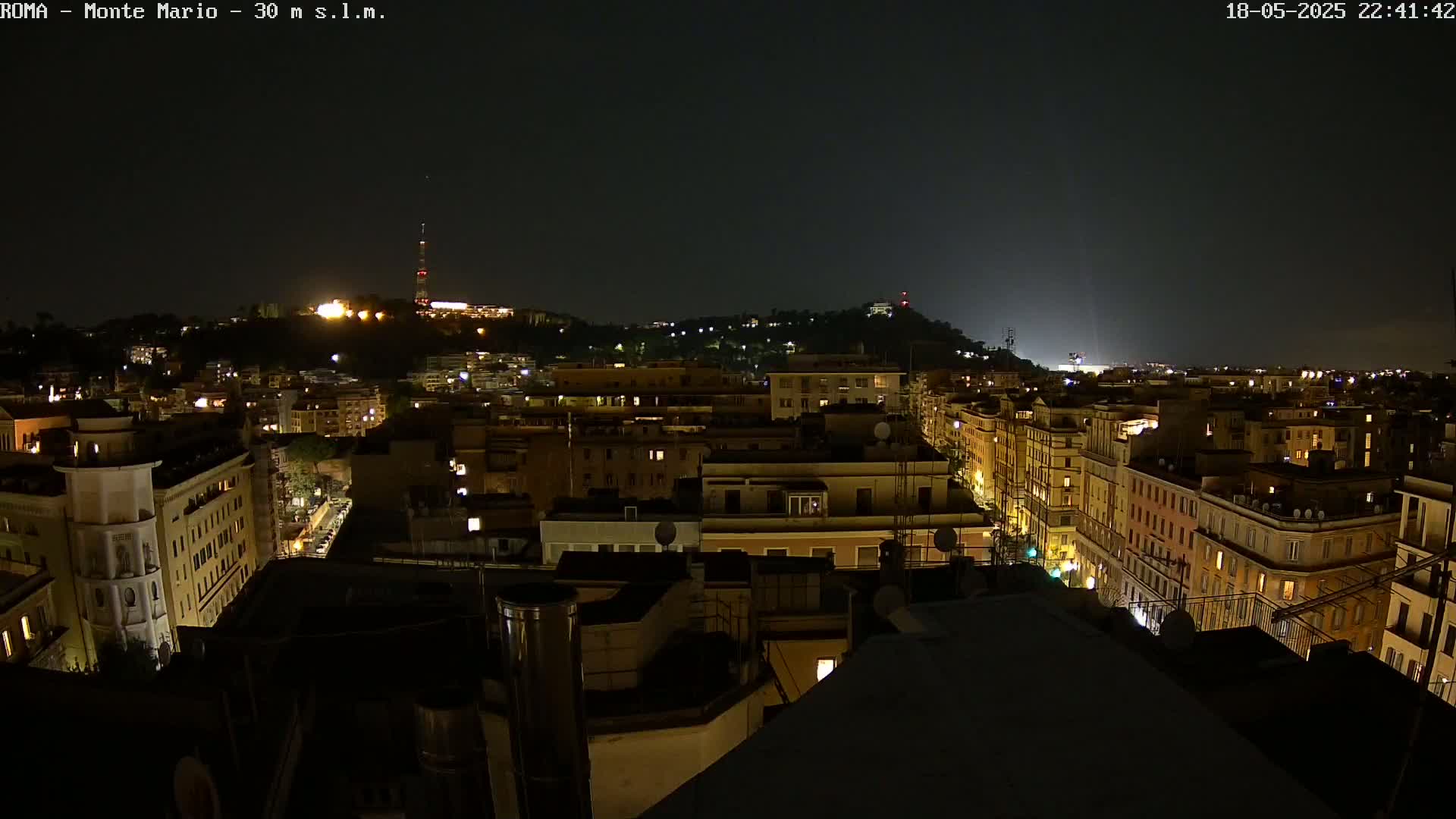 A nighttime view of a city illuminated by streetlights, with a hill in the distance also showing lights.