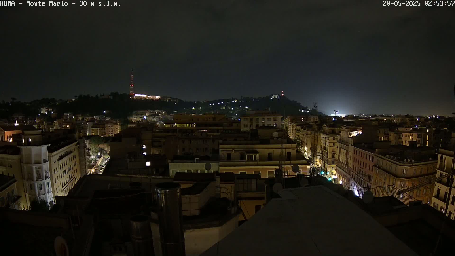A nighttime view of a city with dimly lit buildings, a hill in the background, and a dark, possibly cloudy sky.