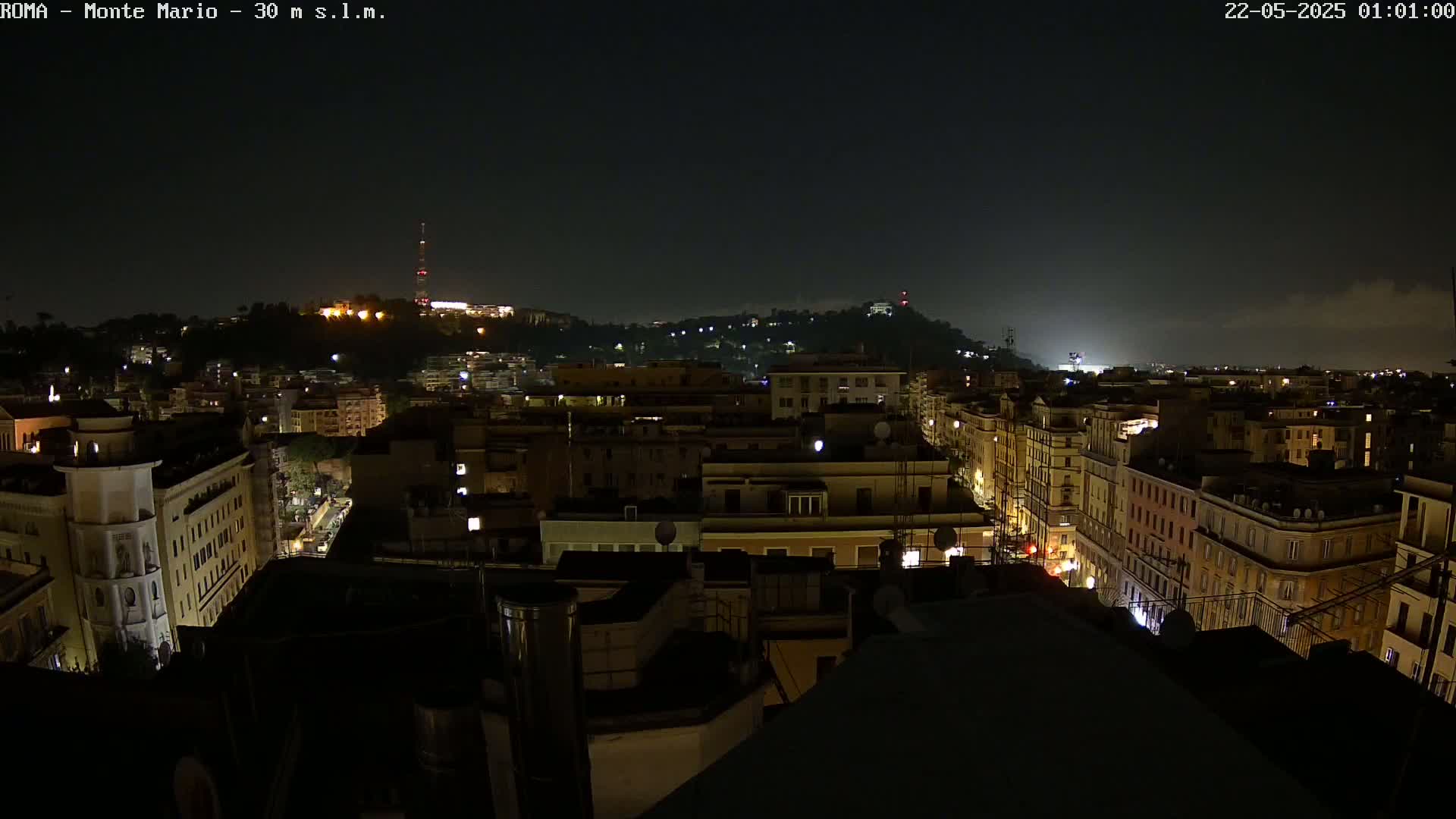 A nighttime view of a city with a hill in the background under a partly cloudy sky.