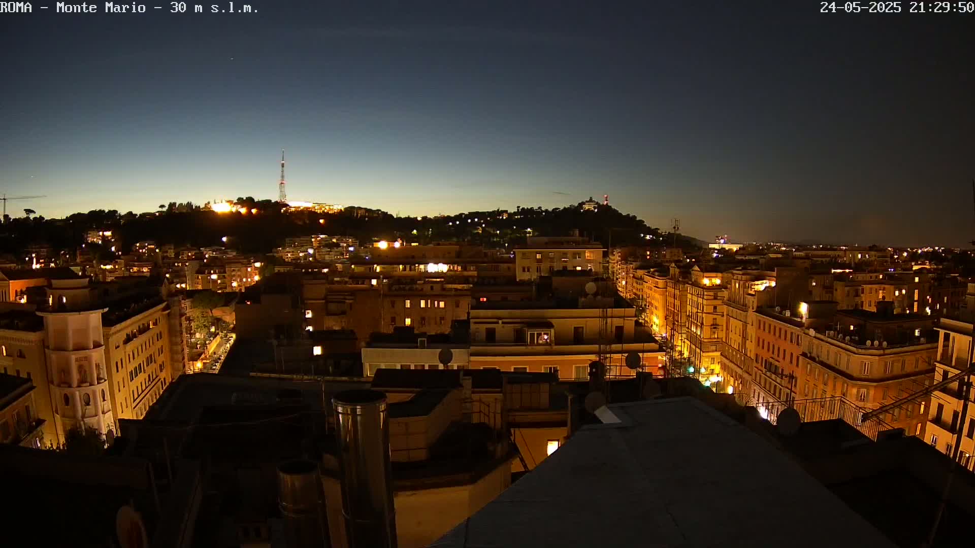 A nighttime view of a city, illuminated by many building lights, with a hill in the background under a clear, dark sky.