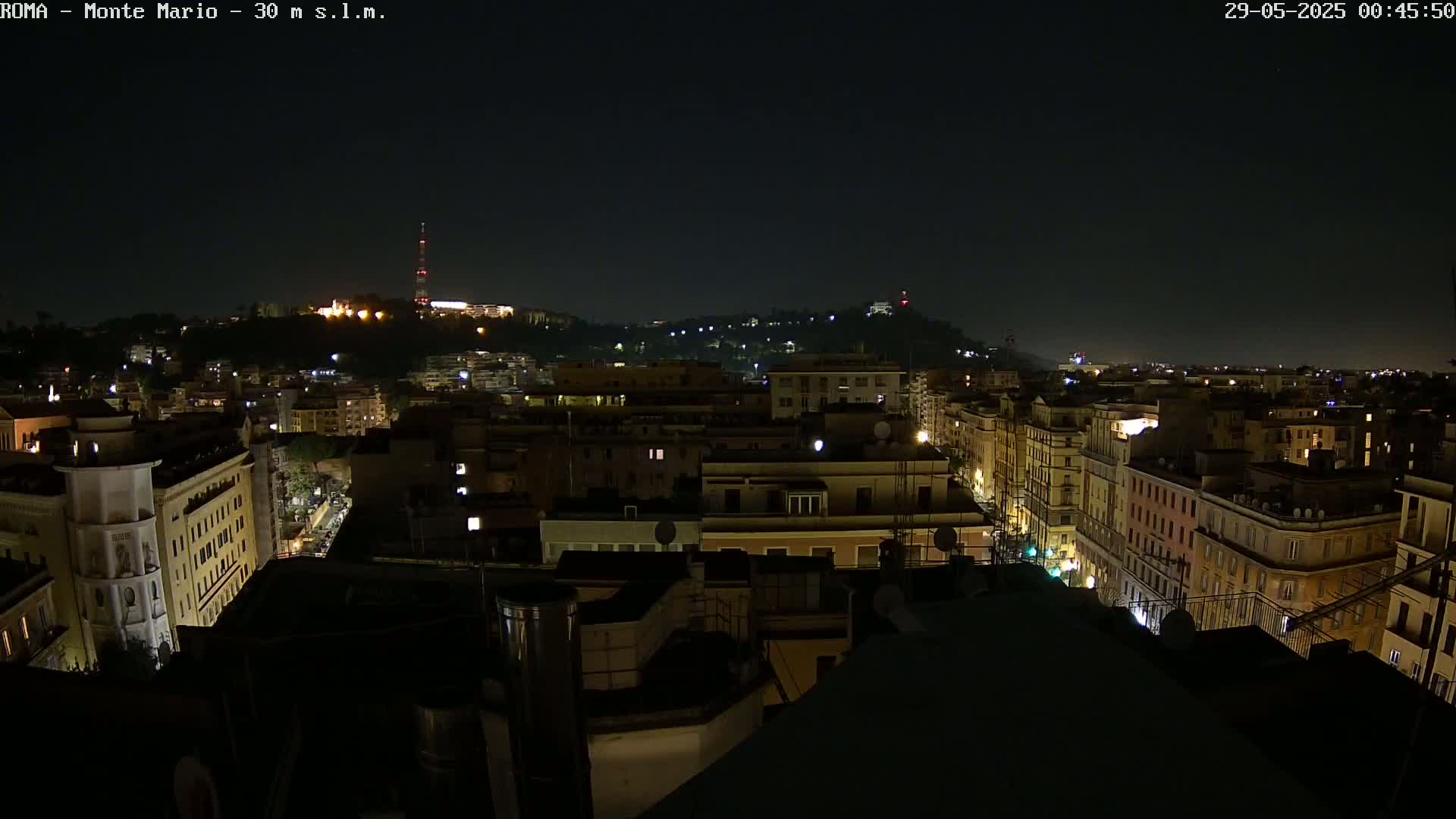A nighttime view of a city, showing many buildings illuminated with streetlights and windows, with a hill in the background under a clear, dark sky.