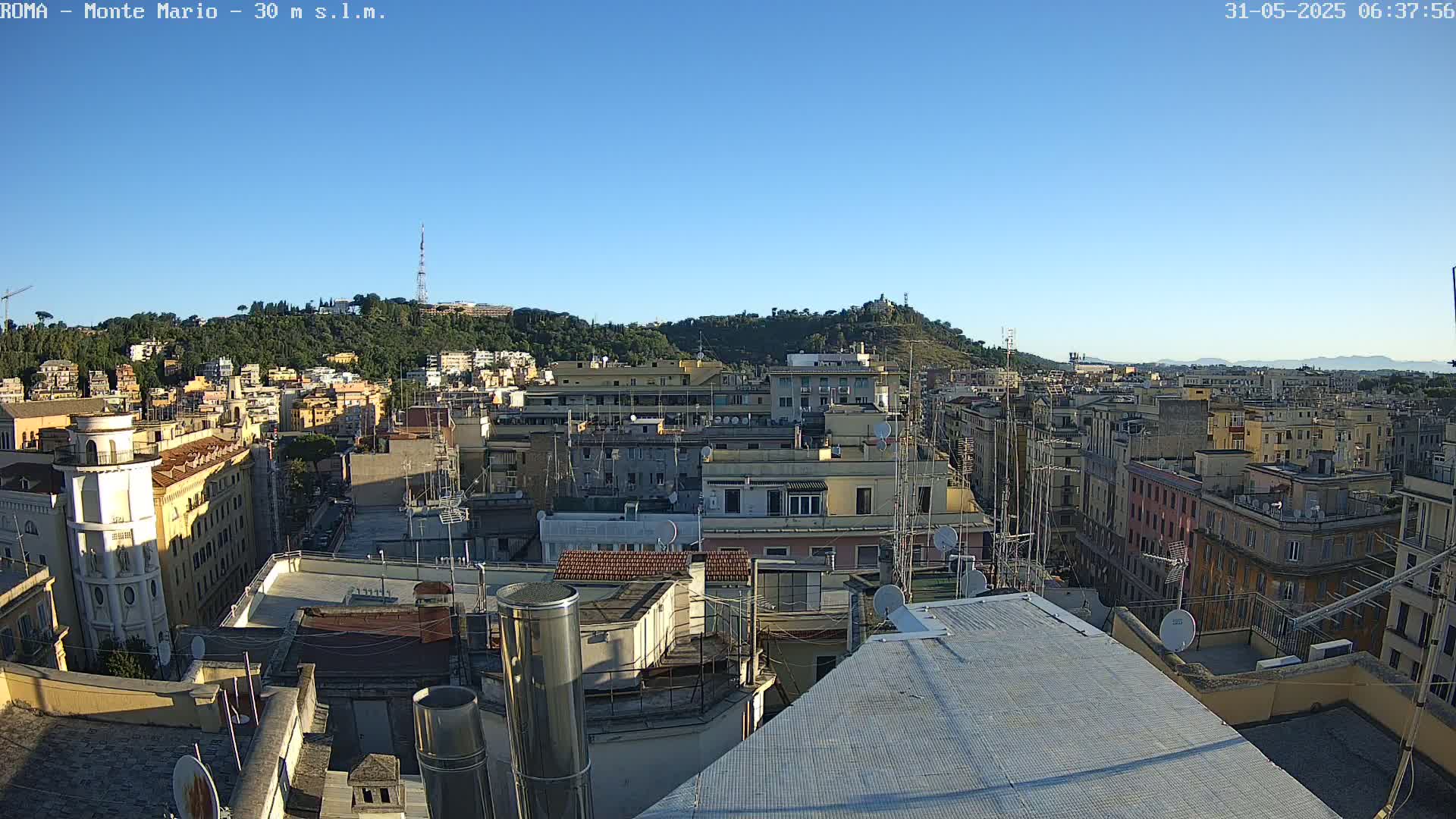 A clear, sunny day reveals a panoramic view of a dense cityscape, with multi-story buildings densely packed together and a hill covered in trees in the background.