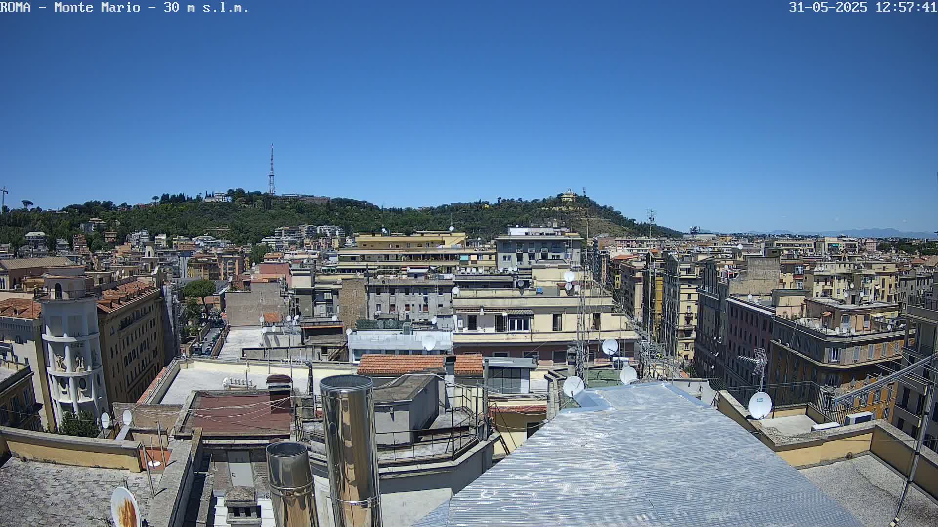 A rooftop view of a densely populated city under a clear blue sky, showing numerous buildings and a hill in the distance.