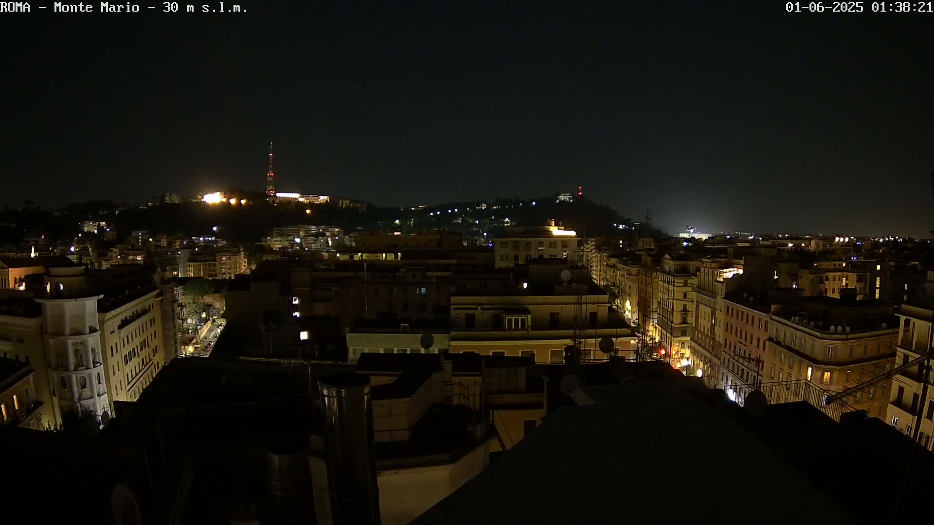A nighttime, high-angle view of a city illuminated by streetlights, with a hill in the distance also showing some lights.