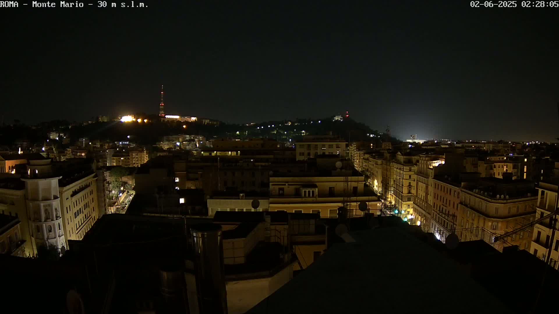 A nighttime aerial view of a city with dimly lit buildings, streets, and a hilltop in the distance under a clear, dark sky.