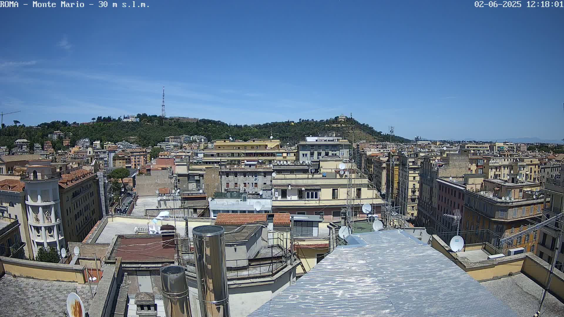 A rooftop view of a densely populated city on a clear, sunny day, showing many buildings extending to a hill in the distance.