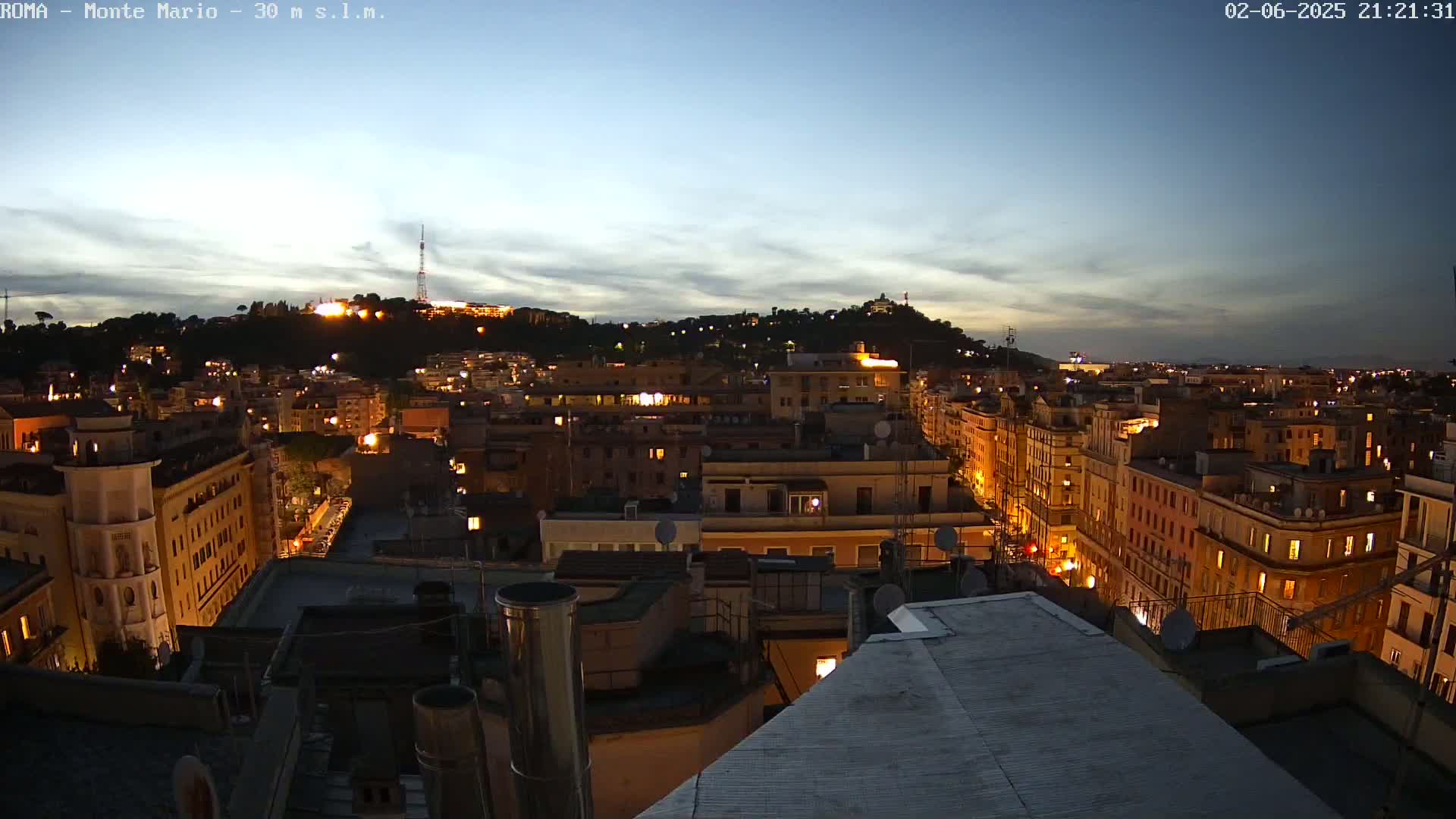 A nighttime view of a city with illuminated buildings, stretching towards a hilltop under a twilight sky.