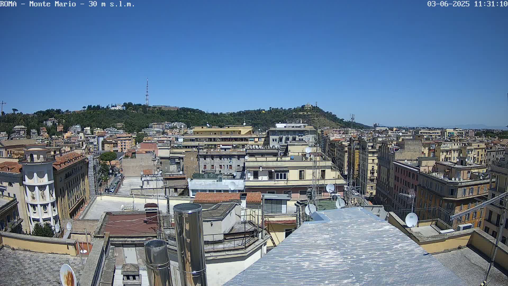A high-angle, sunny-day view of a dense cityscape with numerous buildings, rooftops, and satellite dishes, overlooking a hill in the distance.
