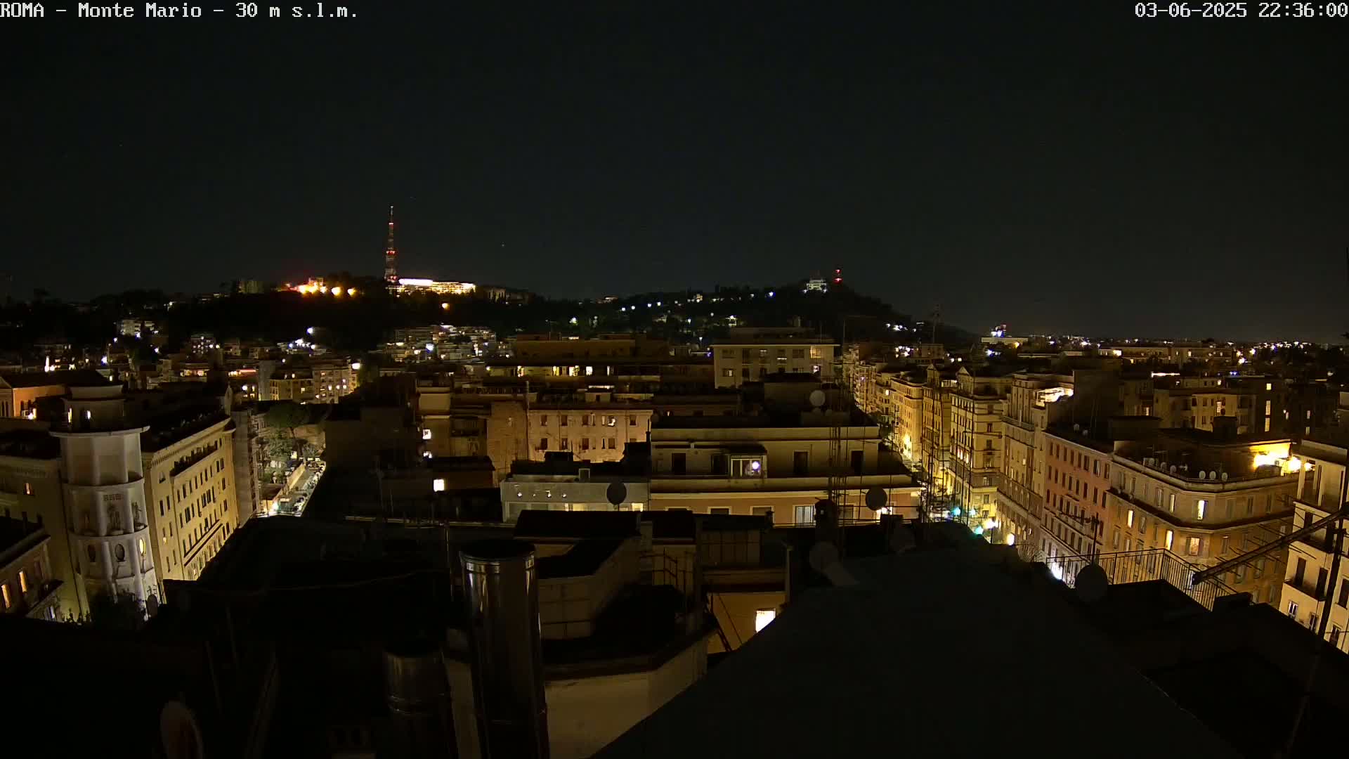 A nighttime view of a city, with illuminated buildings and streets stretching toward a hilltop in the distance under a clear, dark sky.