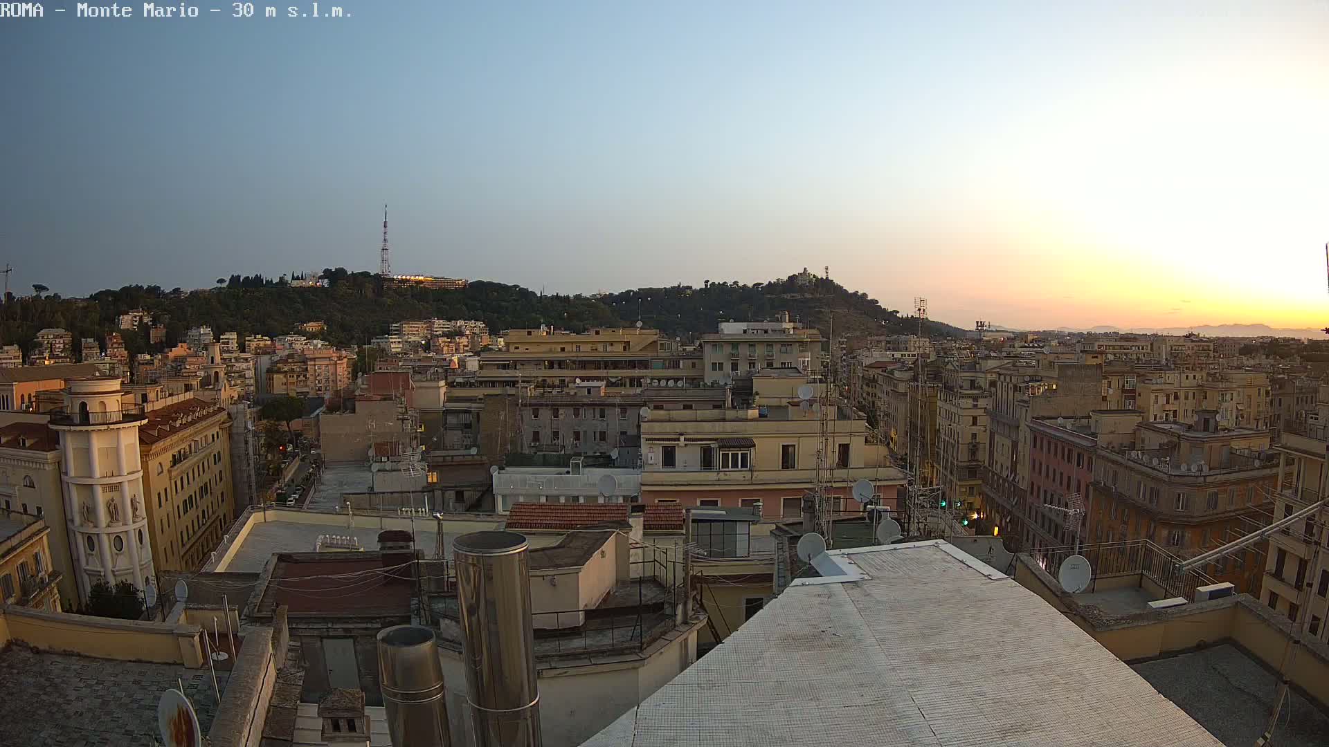 A sunset view shows a cityscape of numerous buildings, with a hill in the background under a clear, pale sky.