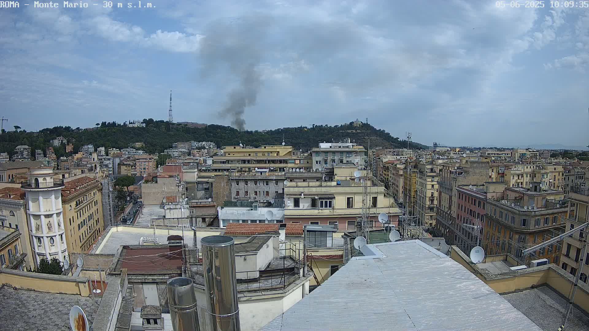 A rooftop view of a city under a partly cloudy sky, with a plume of smoke rising from a hill in the distance.
