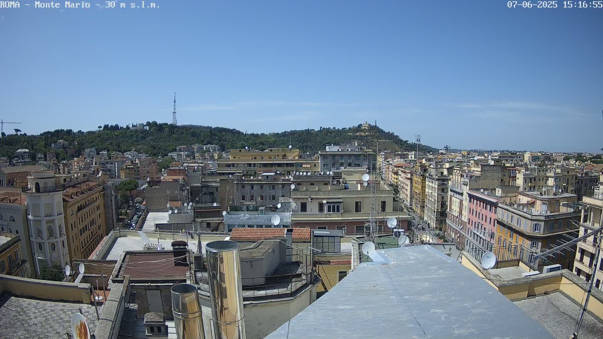 A high-angle, long shot on a clear, sunny day shows a dense cityscape of multi-story buildings, with a hill covered in trees and a tall tower in the distance.