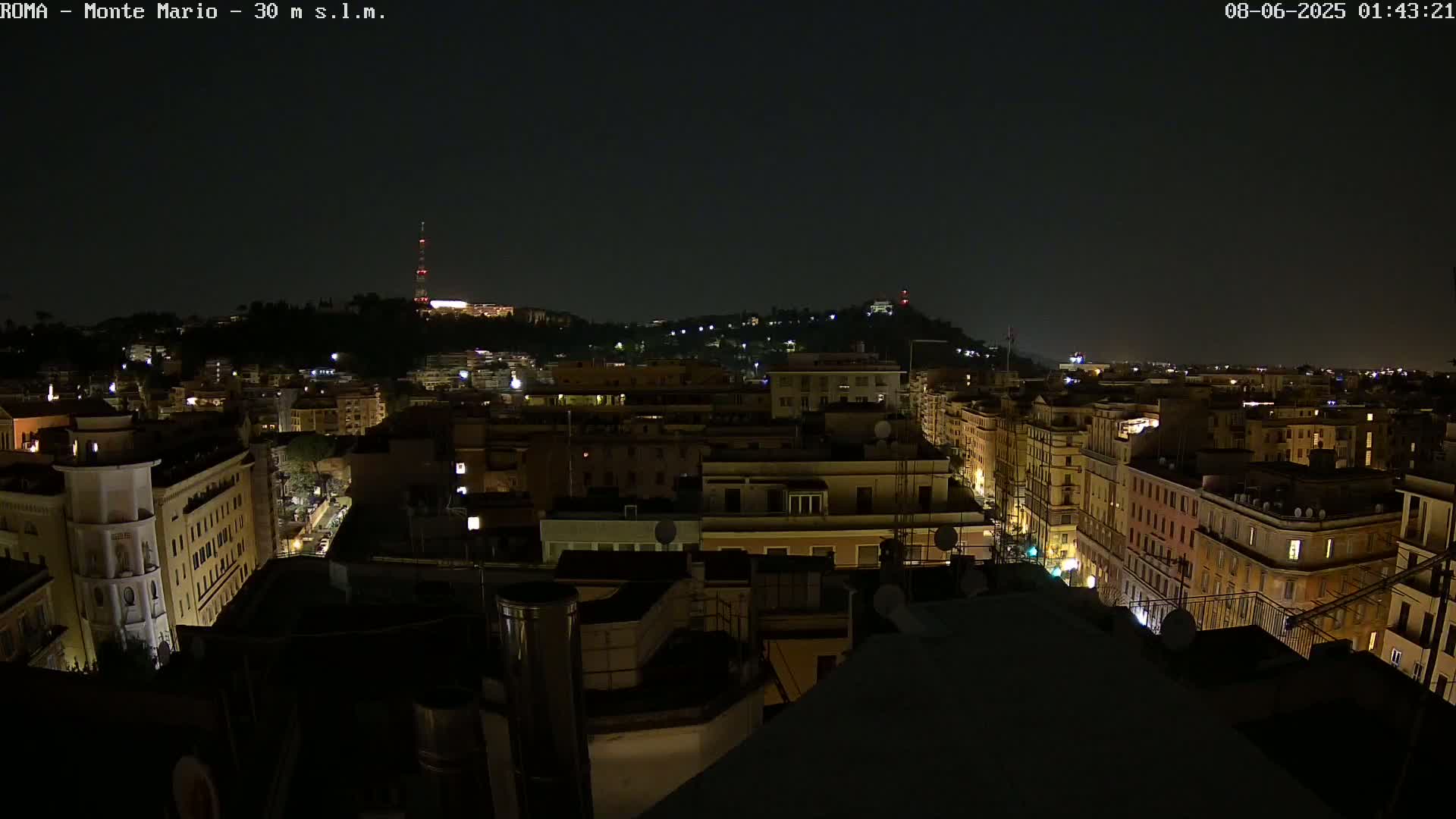 A nighttime view of a city, showing numerous buildings illuminated with amber lights, stretching out towards a hill in the distance under a dark sky.