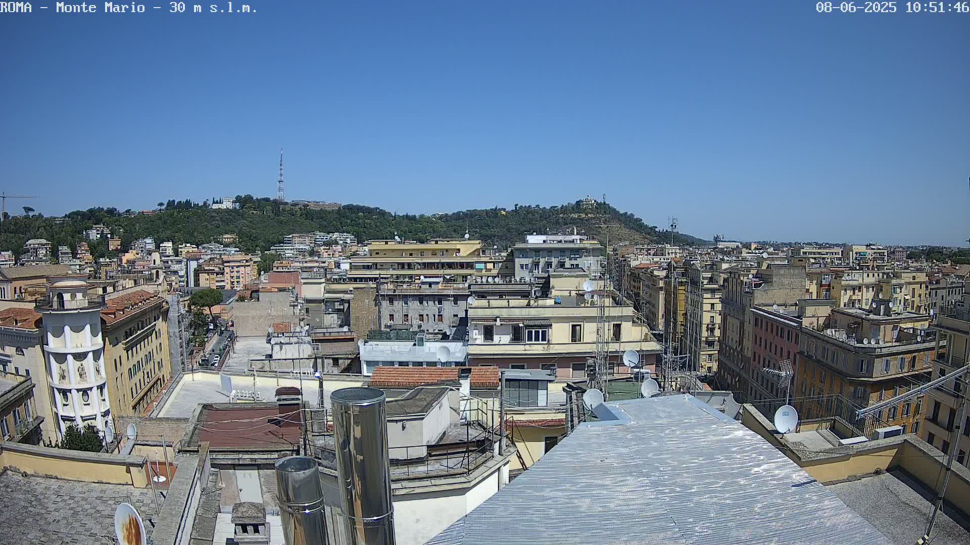 A sunny cityscape view from a rooftop shows numerous buildings extending to a hill in the distance.