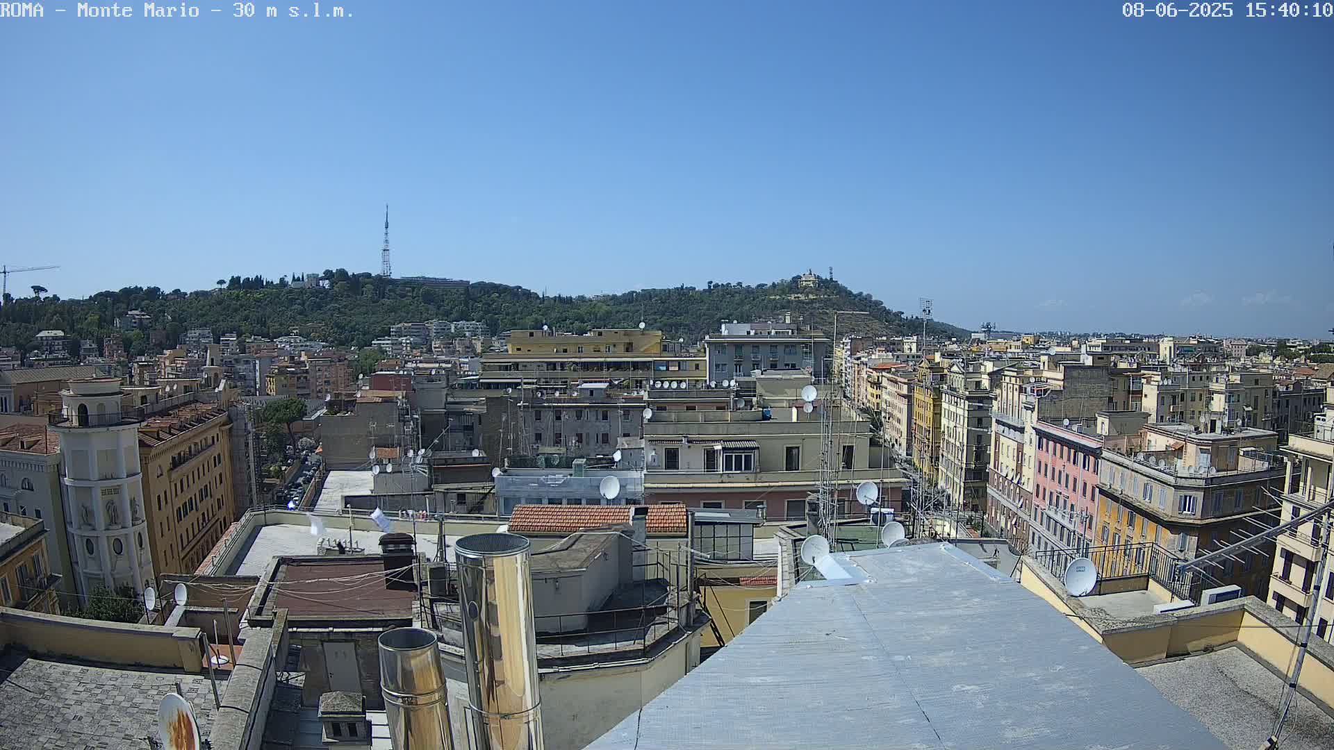 A rooftop view of a densely populated city on a clear, sunny day, showing numerous buildings and a hill in the distance.