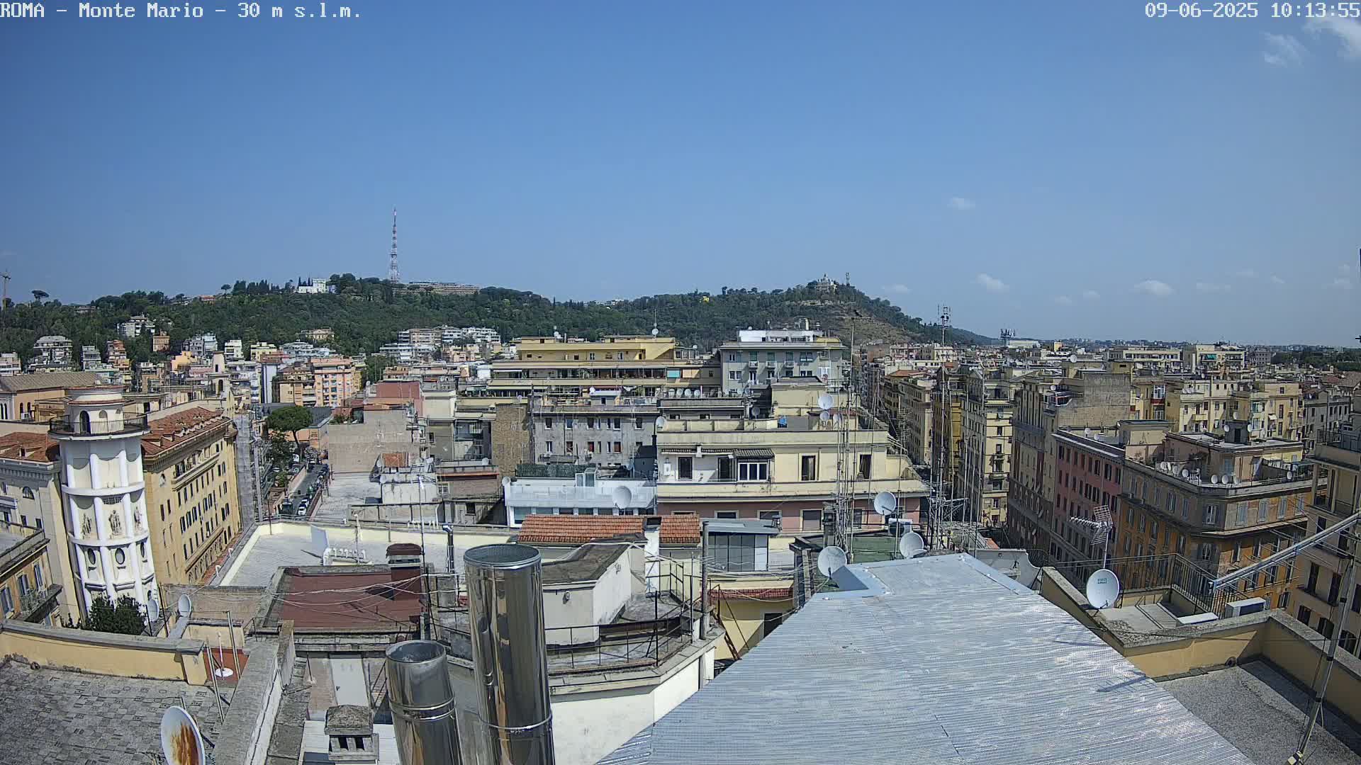 A high-angle, long shot on a clear, sunny day shows a dense cityscape of multi-story buildings, many with rooftop satellite dishes, extending to a distant hill covered in green vegetation.