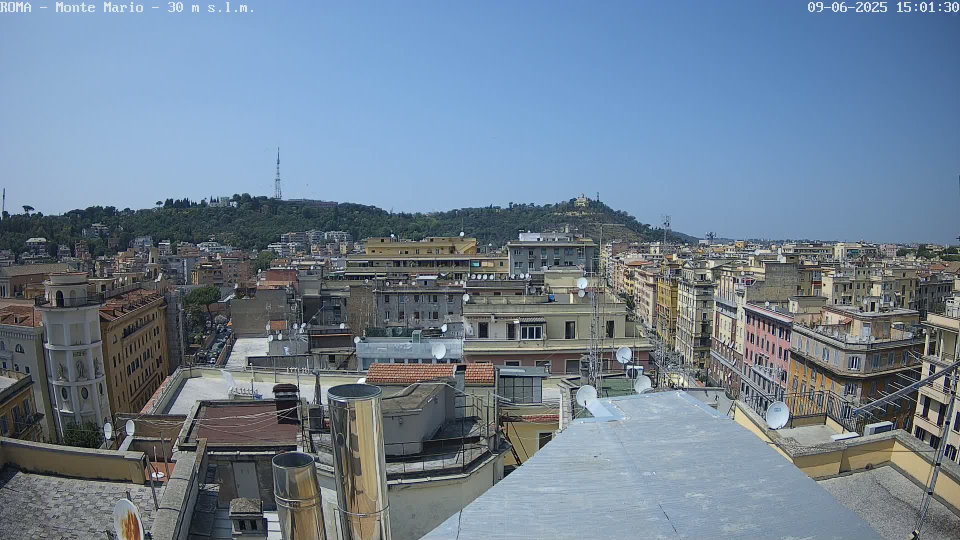A rooftop view of a dense city under a clear blue sky, showing many buildings and a hill in the distance.