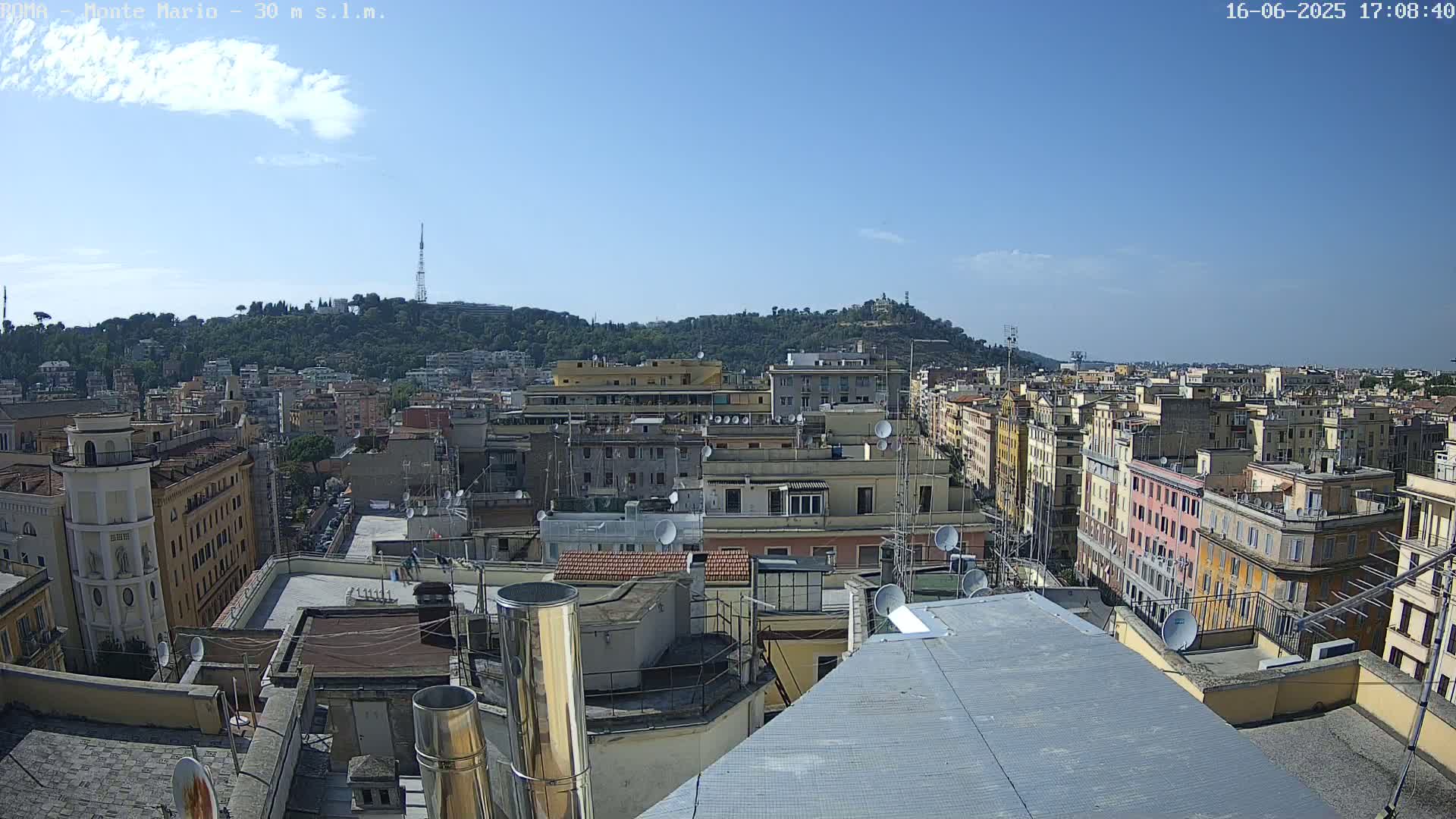 A rooftop view of a city on a clear, sunny day, showing numerous buildings extending to a hill in the distance.