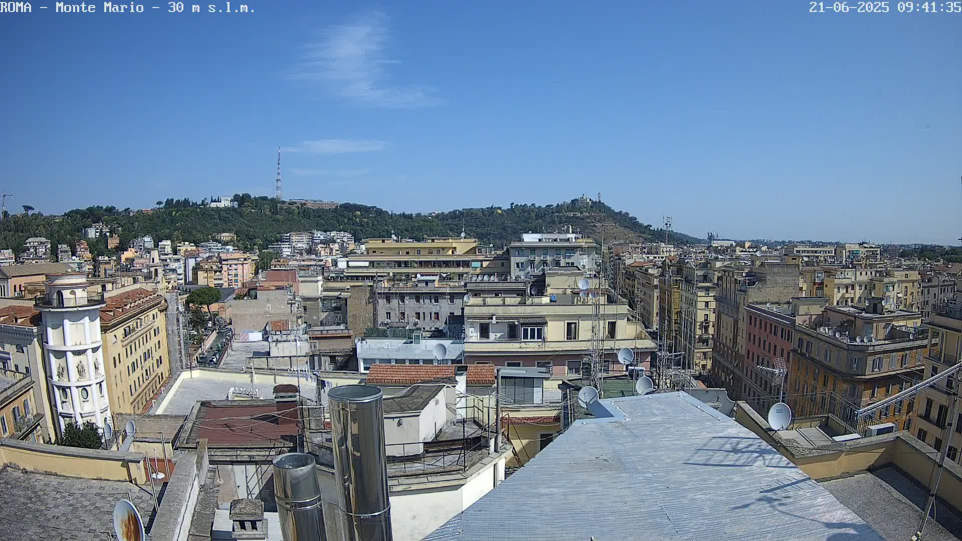 A sunny cityscape view shows many multi-story buildings, rooftops with satellite dishes, and a hill in the background.