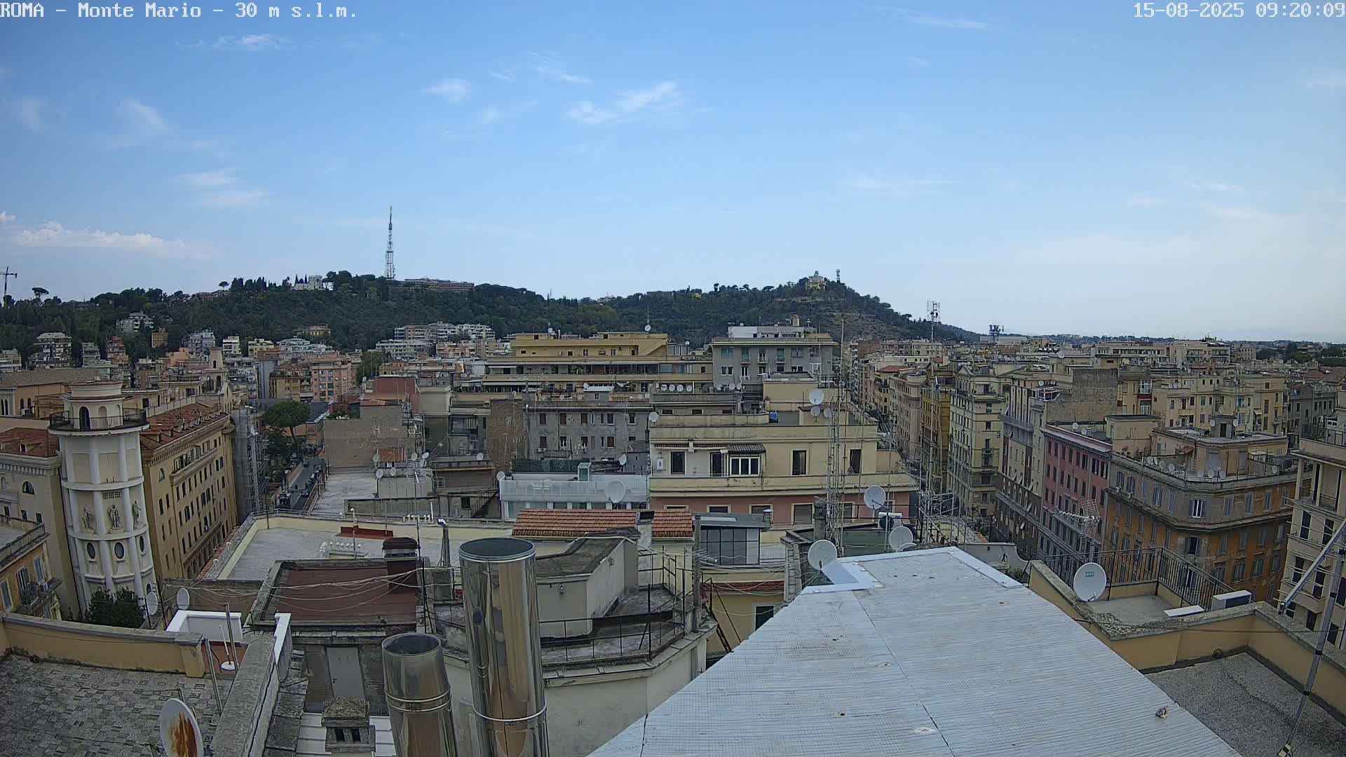 A mostly clear blue sky overlooks a dense cityscape of multi-story buildings, with a tree-covered hill in the background.