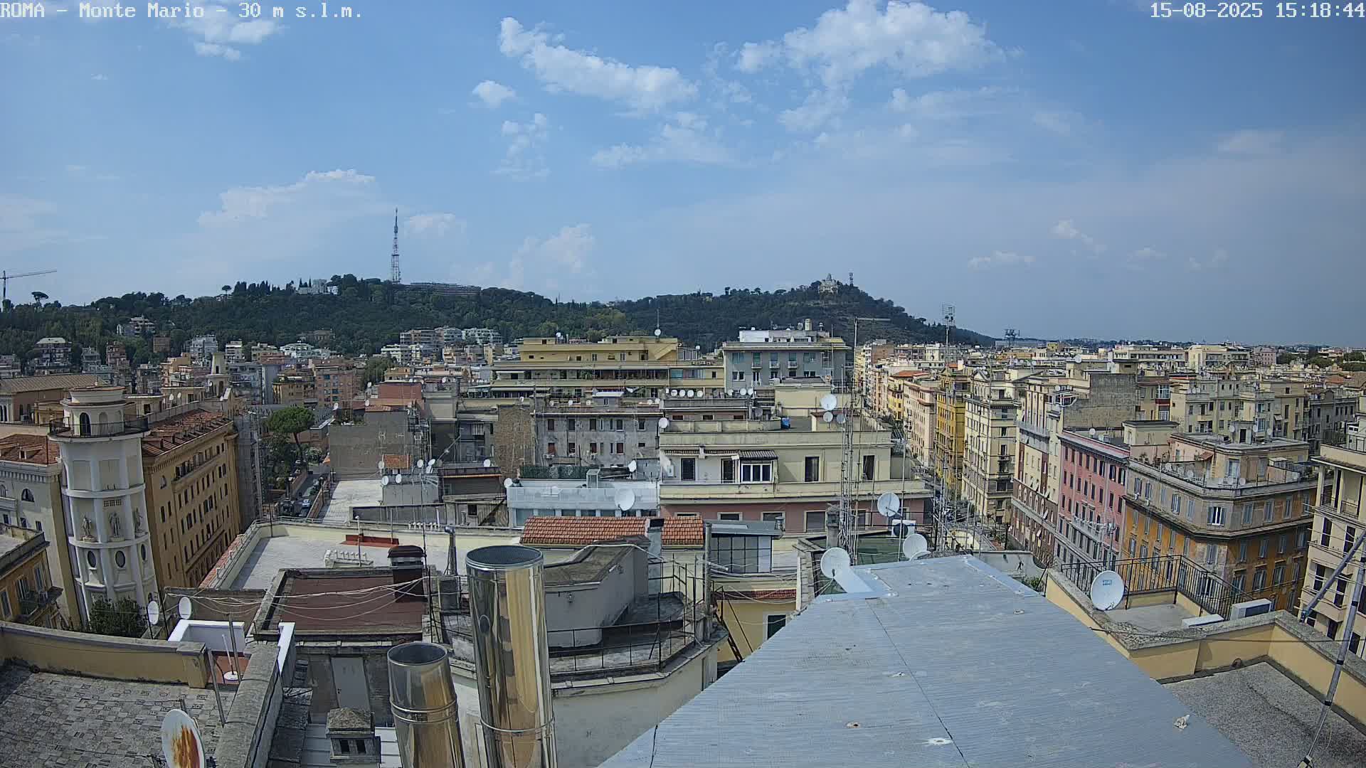 A mostly clear blue sky overlooks a dense cityscape of multi-story buildings, with a tree-covered hill in the background.