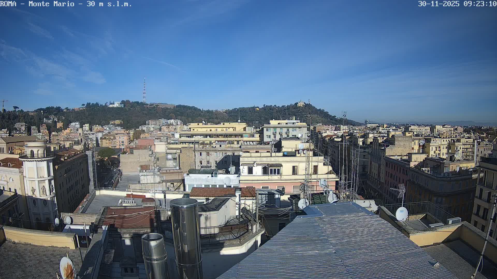 A panoramic view of a densely packed European city unfolds from a high vantage point, revealing numerous rooftops, multi-story buildings, and a distant tree-covered hill topped with an antenna tower, all bathed in bright sunlight under a clear blue sky.