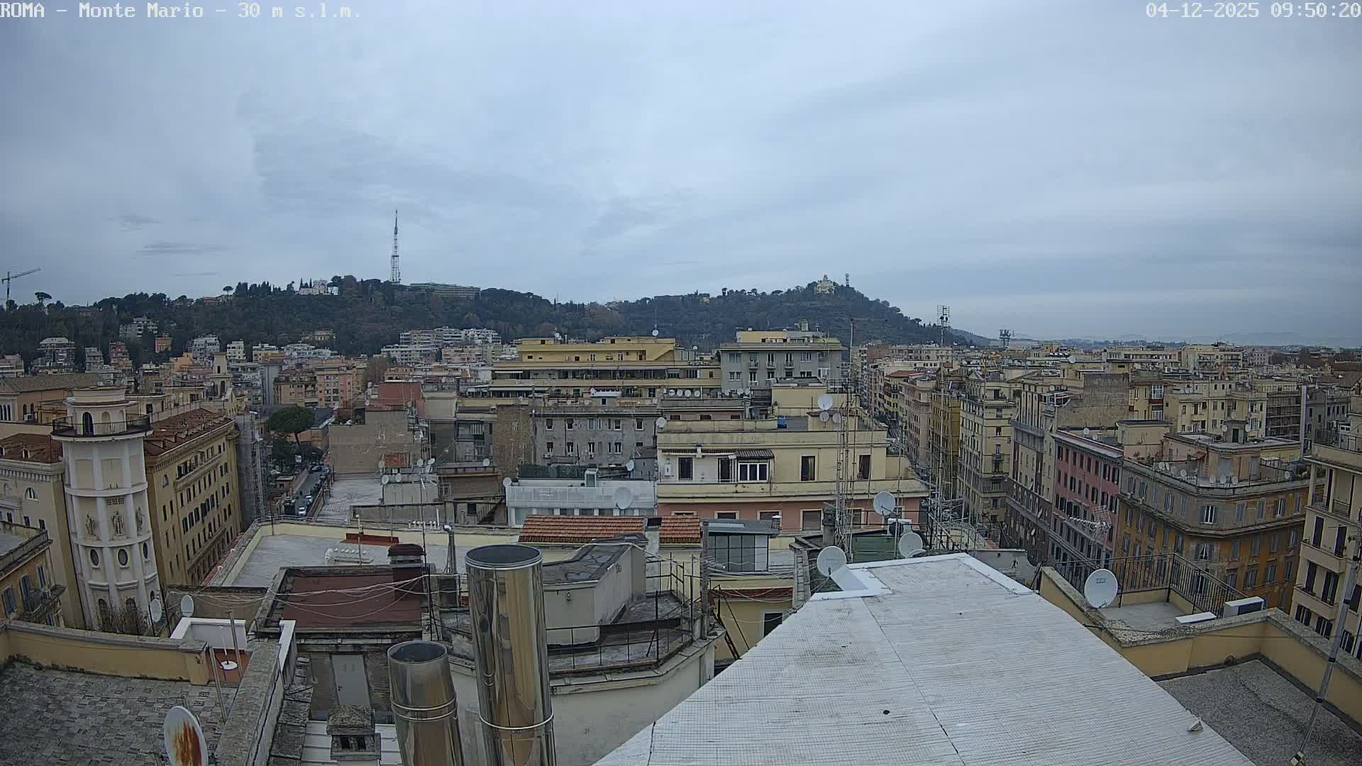 An overcast and cloudy sky looms over a panoramic view of a densely packed city from a high vantage point, revealing numerous rooftops in the foreground and urban buildings stretching towards tree-covered hills in the distance.