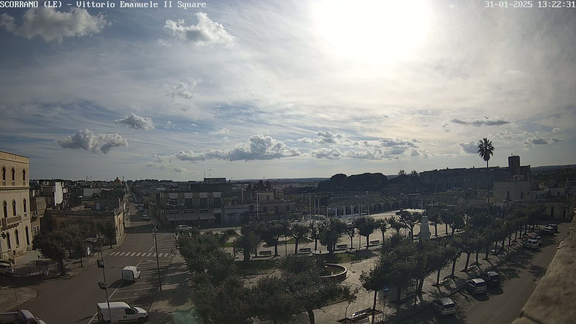 A partly sunny day reveals a town square with a tree-lined pathway, buildings lining a street, and vehicles parked along the road, under a sky filled with scattered clouds.